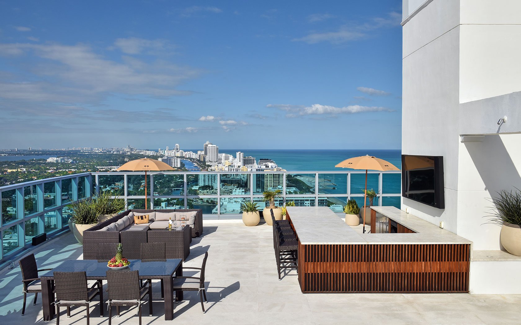 Rooftop patio overlooking a cityscape and ocean, featuring outdoor seating, dining table, parasols, and a built-in bar with a television.