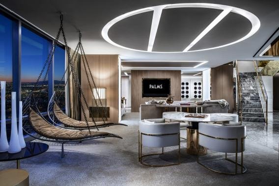 Luxury hotel suite with modern design, featuring two hanging chairs, a round marble dining table with chairs, a TV screen, and large windows showing city views.