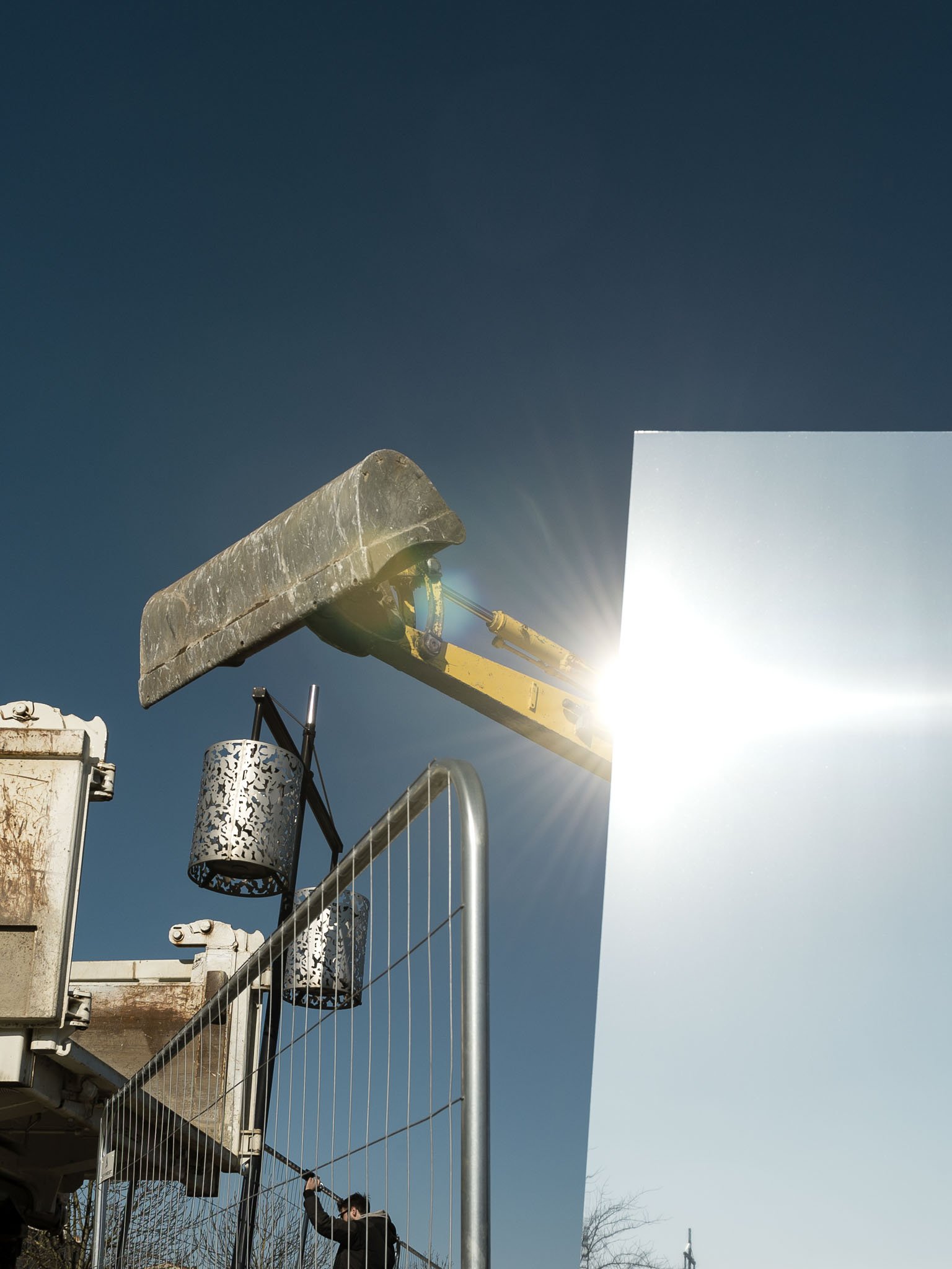 Scénographie en extérieur pour un chantier dans l'espace public. Détail chantier avec une grue de construction derrière un grand panneau miroir, sur fond de ciel bleu.