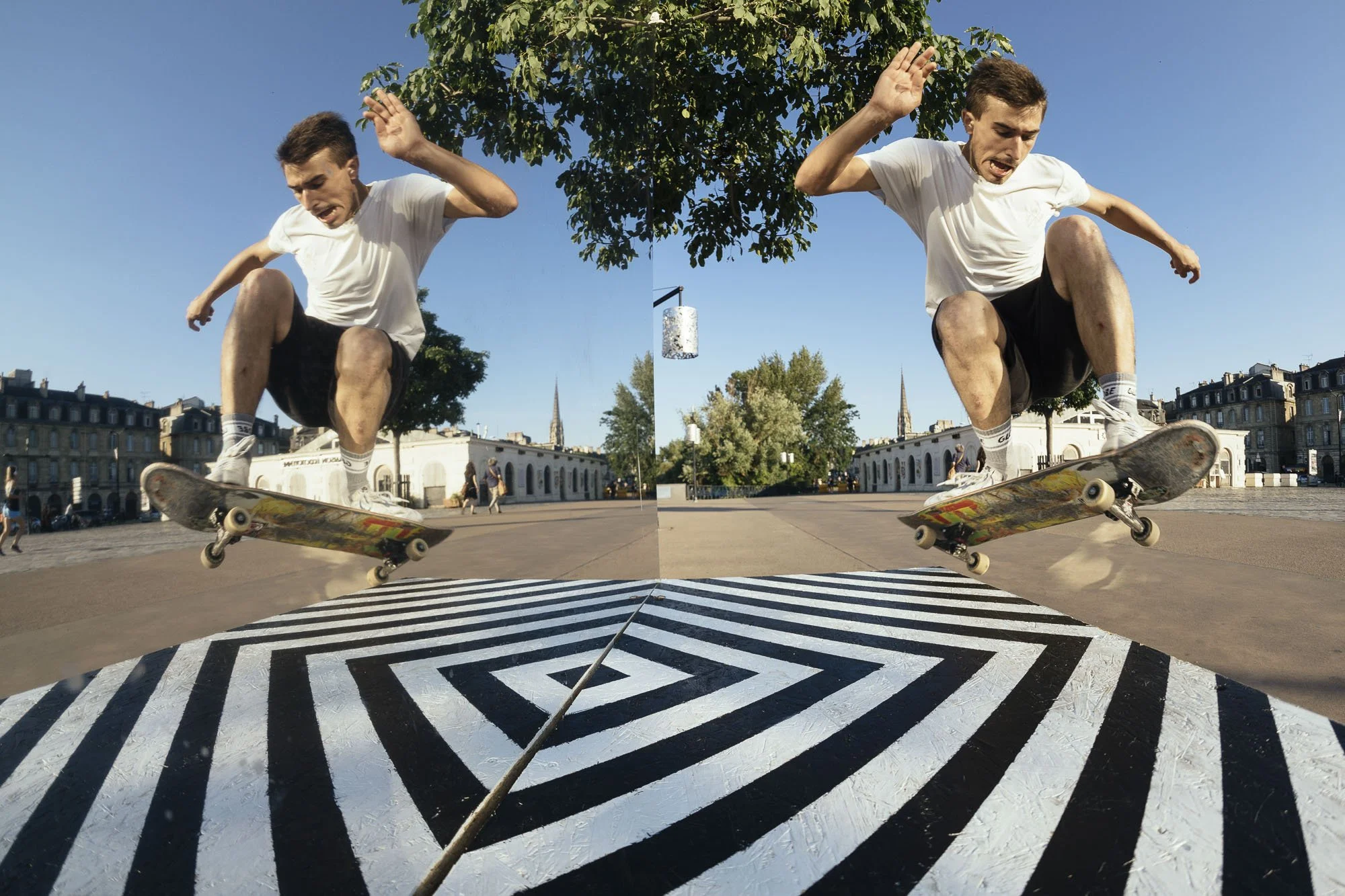 Scénographie en extérieur pour un chantier dans l'espace public. Un jeune homme et son reflet sur le miroir, en train de faire une figure de skateboard dans une zone urbaine en plein air, avec un ciel bleu clair et des bâtiments en arrière-plan.