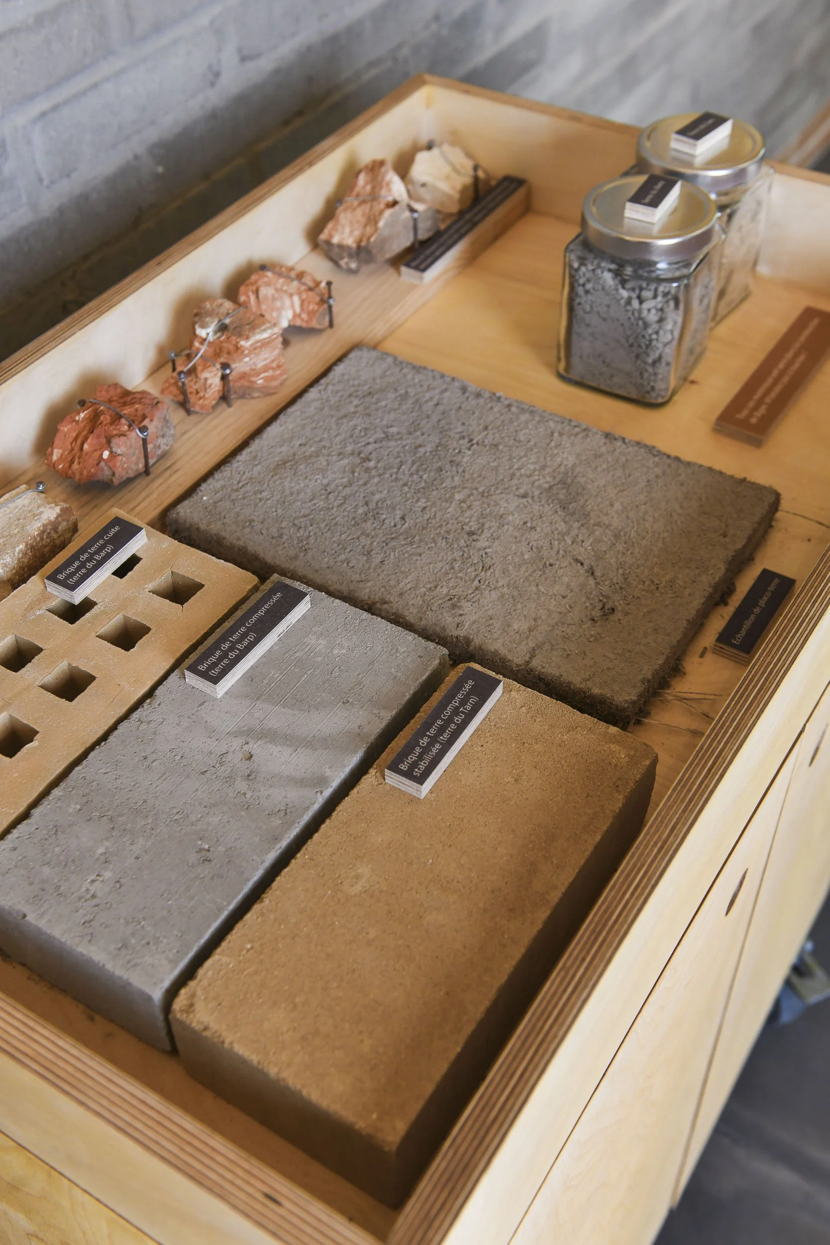 Scénographie d'exposition. Échantillons de briques et matériaux de construction en terre crue exposés sur une table en bois
