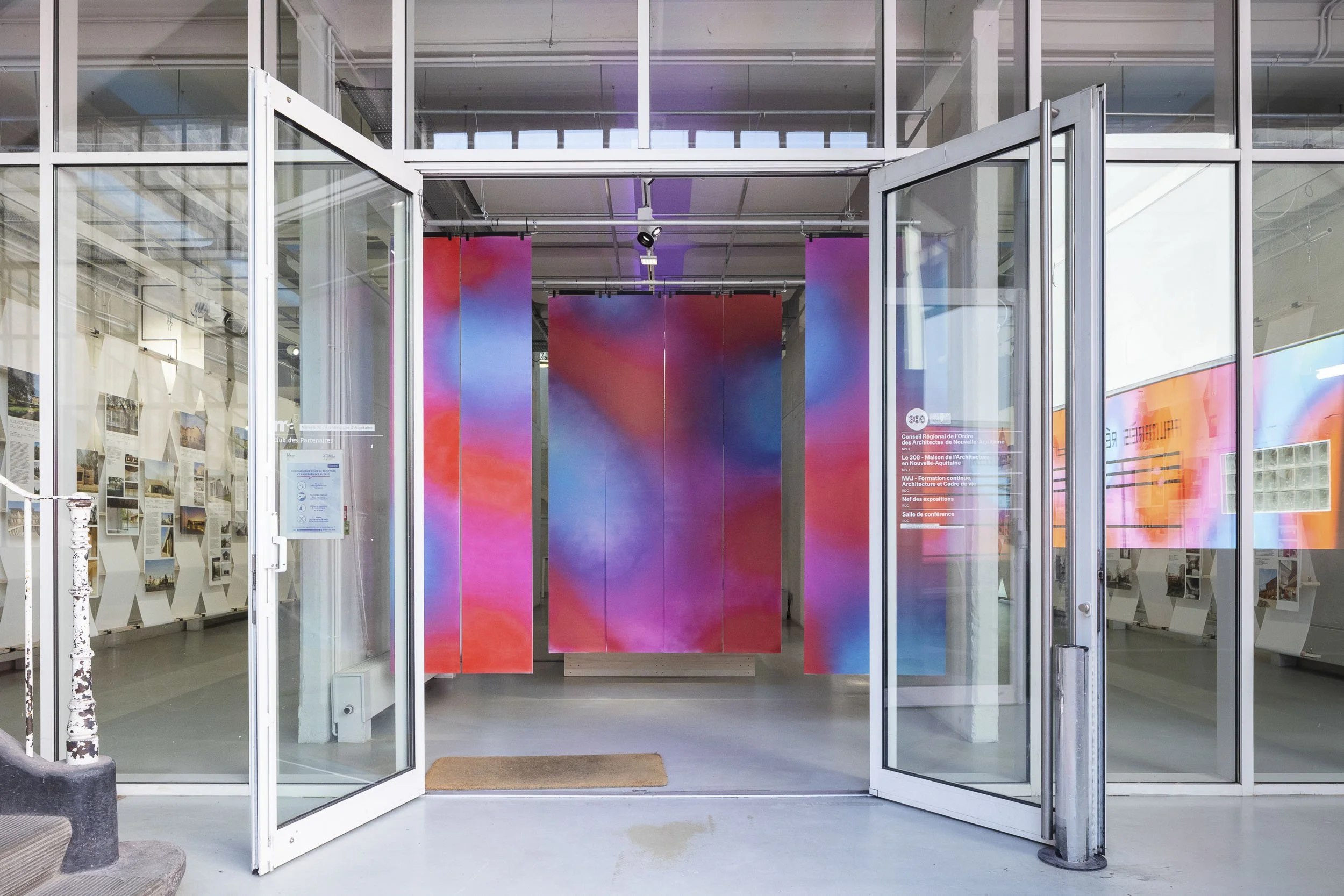 Scénographie d'exposition. Vue sur l'entrée d'une exposition d'architecture avec une façade vitrée, et des motifs colorés abstraits derrière, et plusieurs panneaux d'informations ou de photos sur les côtés dans un espace d'exposition.