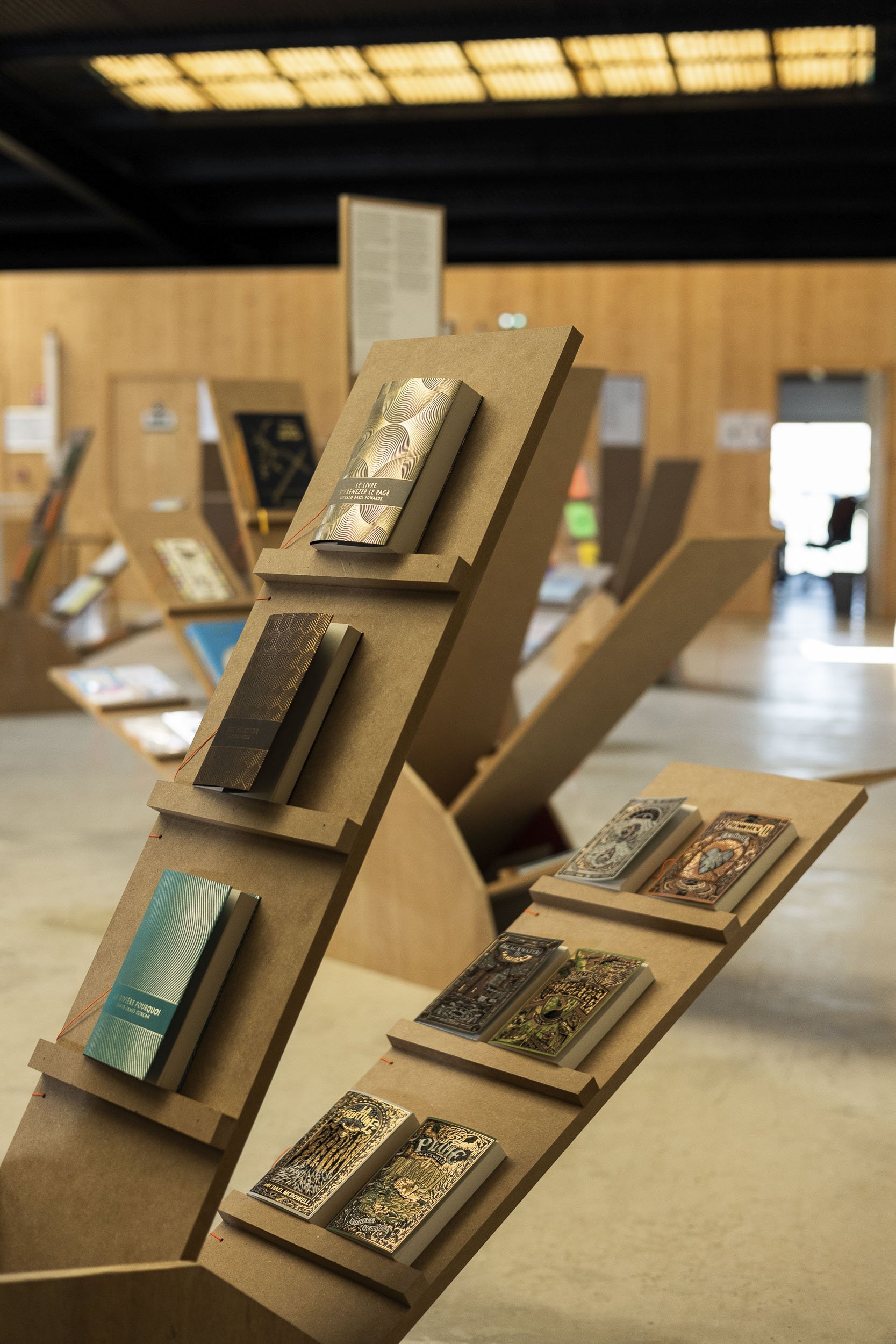 Scénographie d'exposition en intérieur, présentant des livres graphiques. Vue de détail sur le mobilier scénique d'exposition, des grands modules en bois en forme d'éventail, sur lesquels sont posés les livres, ouvrages.