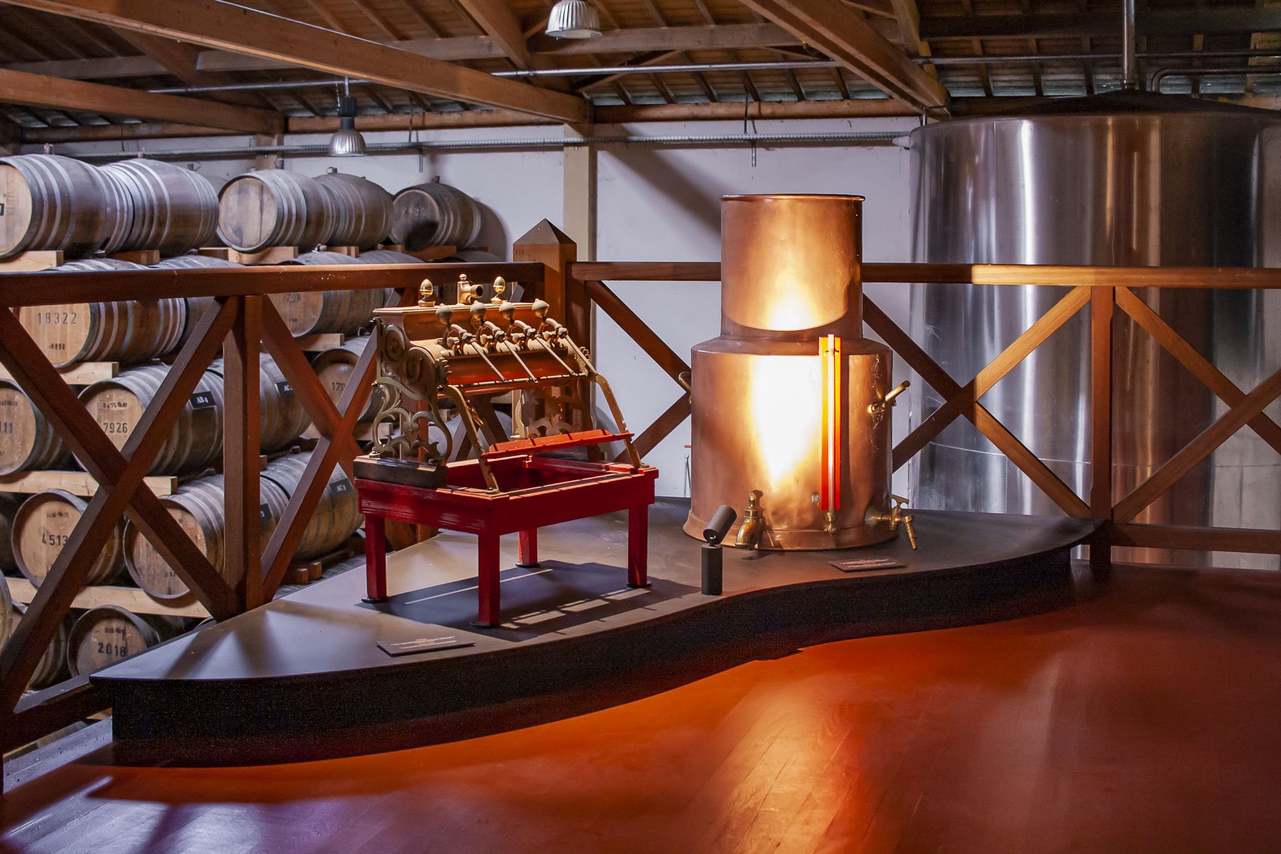 Scénographie d'exposition. valorisation du savoir faire. Intérieur d'un chai à spiritueux, avec des tonneaux en chêne, du contenu d'exposition, des grandes photos, des vieux outils, des plateformes en bois peintes en noir avec des courbes, au sol.
