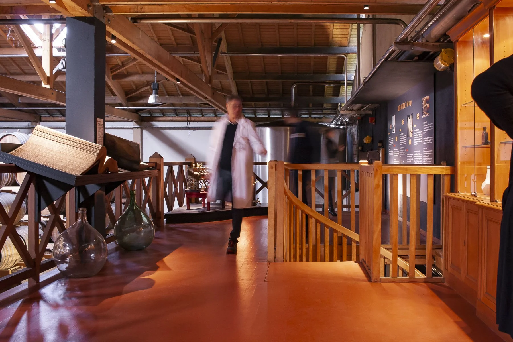 Scénographie d'exposition. Intérieur d'un chai à spiritueux, avec des tonneaux en chêne, du contenu d'exposition, des grandes photos, des vieux outils, des plateformes en bois peintes en noir avec des courbes, au sol, et une personne se promenant.