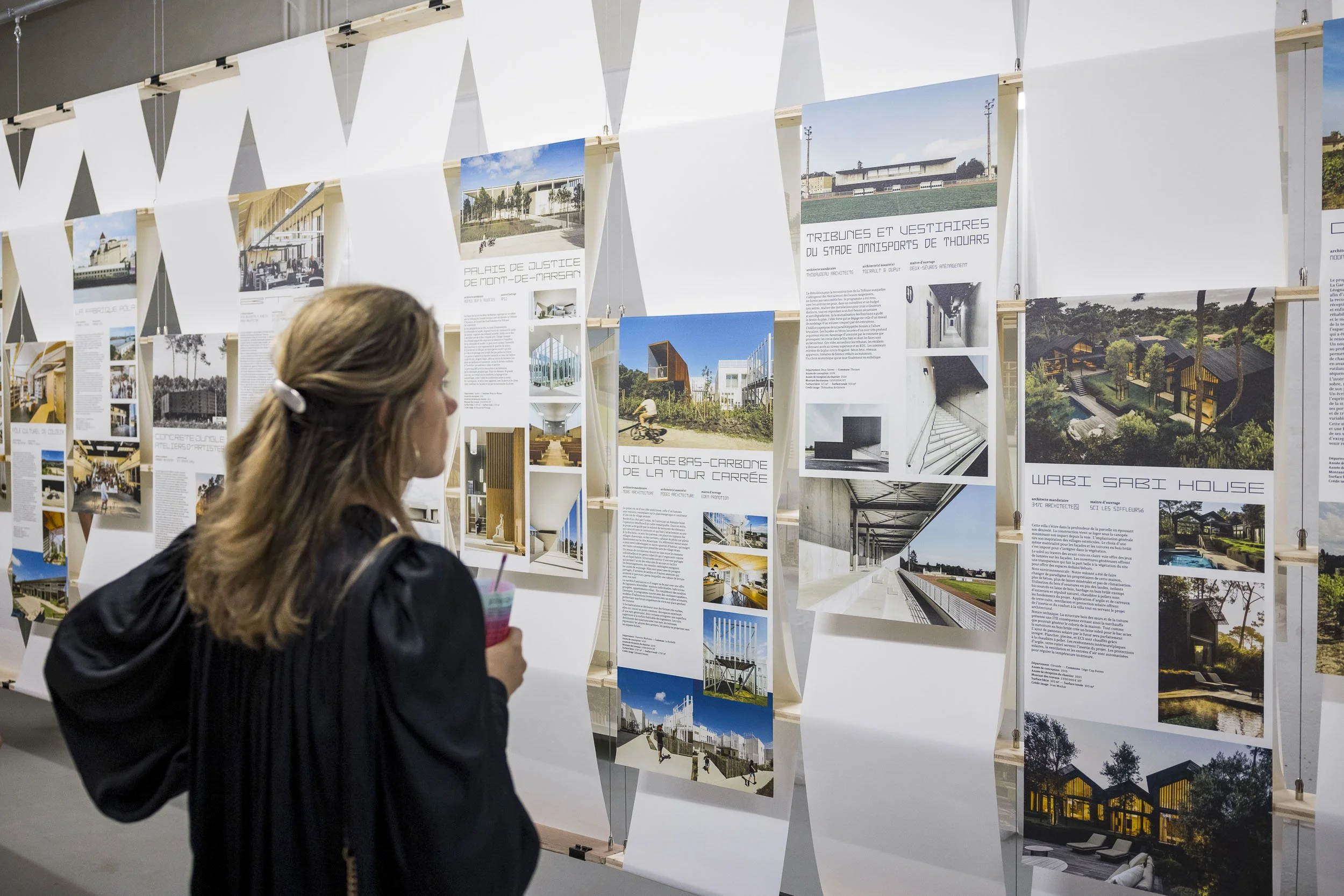 Scénographie d'exposition. Jeune femme regardant une exposition de projets d'architectures, habitation, public, tertiaire, sur de longs kakémonos en papier imprimés.