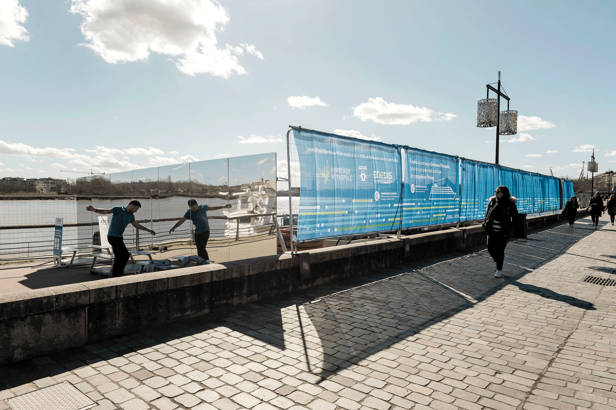 Scénographie en extérieur pour un chantier dans l'espace public. et un habillage de chantier singulier, avec une façade en grands panneaux miroirs et des bâches bleu avec un effet drapé. Des gens et des bâtiments au cœur de la ville, à Bordeaux.