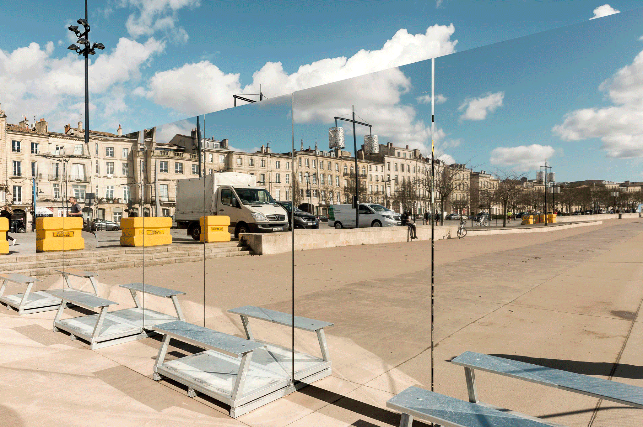 Scénographie en extérieur pour un chantier dans l'espace public. Reflet d'un bâtiment urbain sur des miroirs géants dans un espace public ensoleillé, avec des bancs en métal, des voitures, et un ciel bleu avec des nuages.