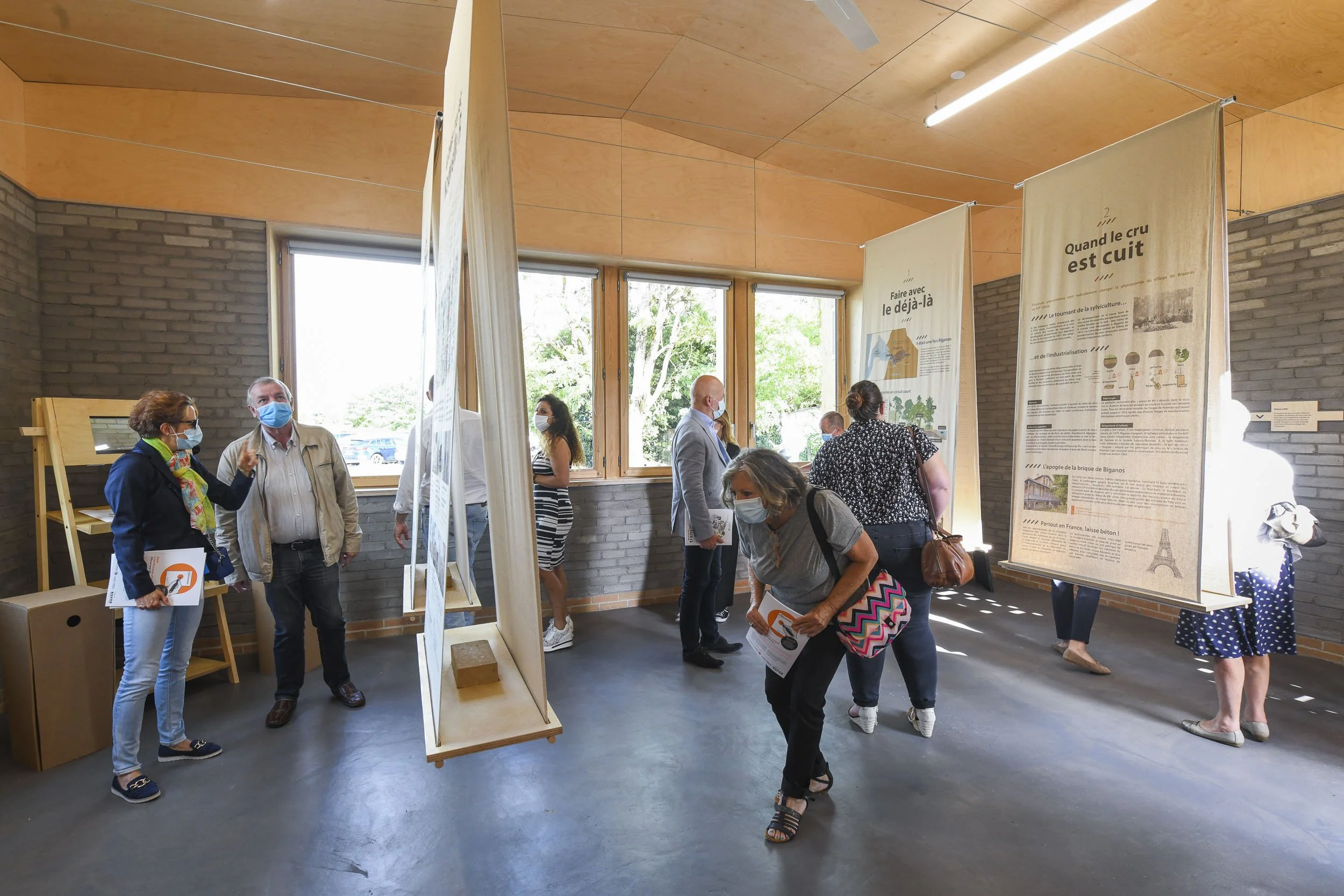 Scénographie d'exposition. Plusieurs personnes, regardant des panneaux d'information suspendus ou debout. La pièce a un mur en briques de terre crue grise, un plafond en bois clair, et de grandes fenêtres laissant entrer la lumière naturelle.
