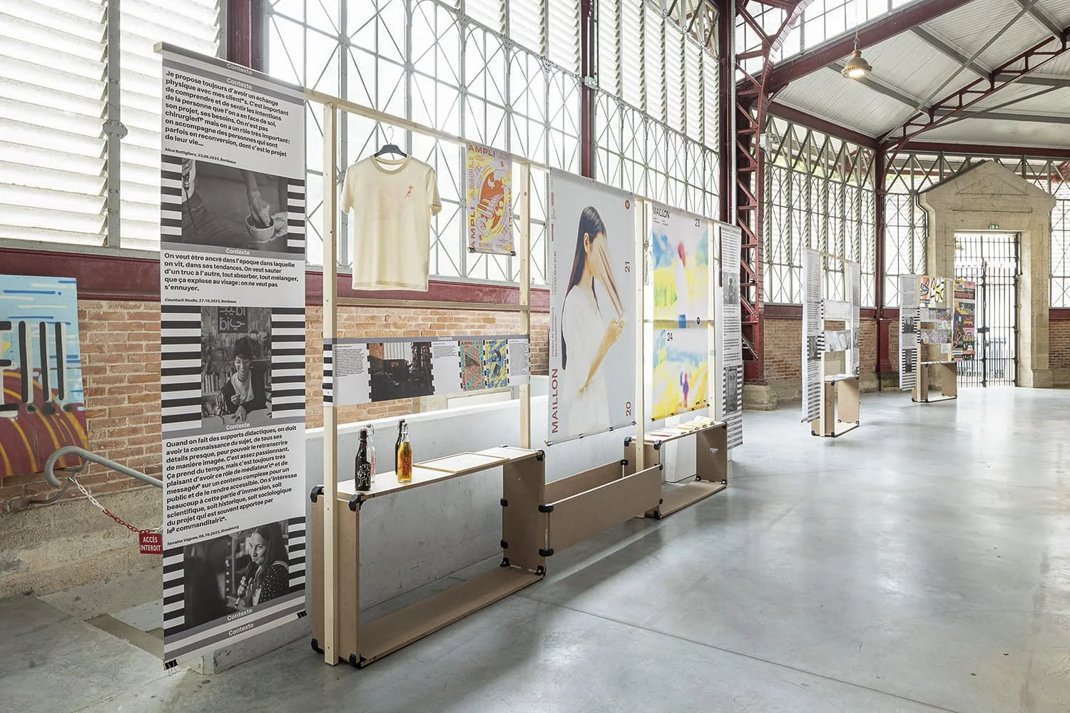 Scénographie d'exposition en intérieur, présentant des projets graphiques. Dans une grande galerie avec de grandes fenêtres en verre et des affiches suspendues sur des mobiliers d'exposition en bois.