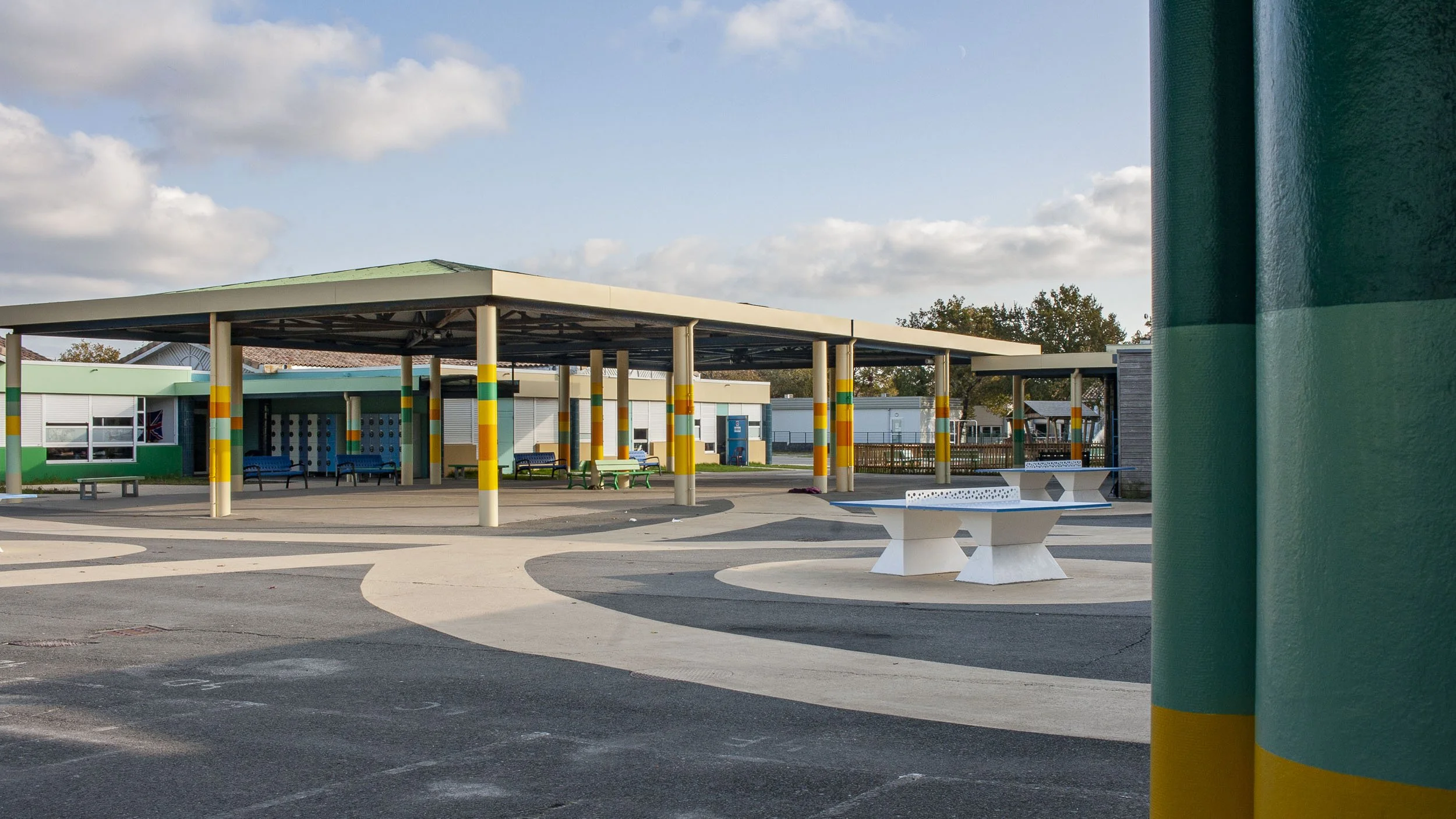 Identité des lieux, requalification d'une cour dans un collège. Aperçu de la cour rénovée, avec des colonnes peintes en plusieurs couleurs propres à l'identité du collège, des jolis cheminements au sol en beige sable, et des tables de ping-pong.