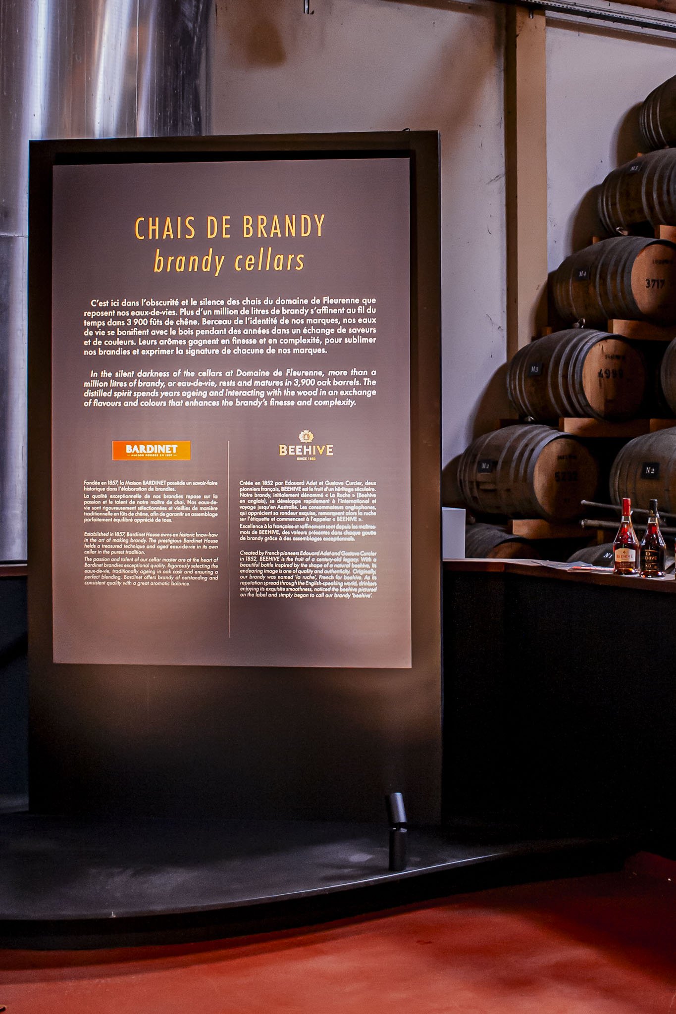 Scénographie d'exposition et valorisation de l'outil de production. Un panneau d'information sur les chais de brandy, avec des descriptions en français et anglais sur l'histoire et la fabrication, placé à l'entrée du chai de spiritueux.