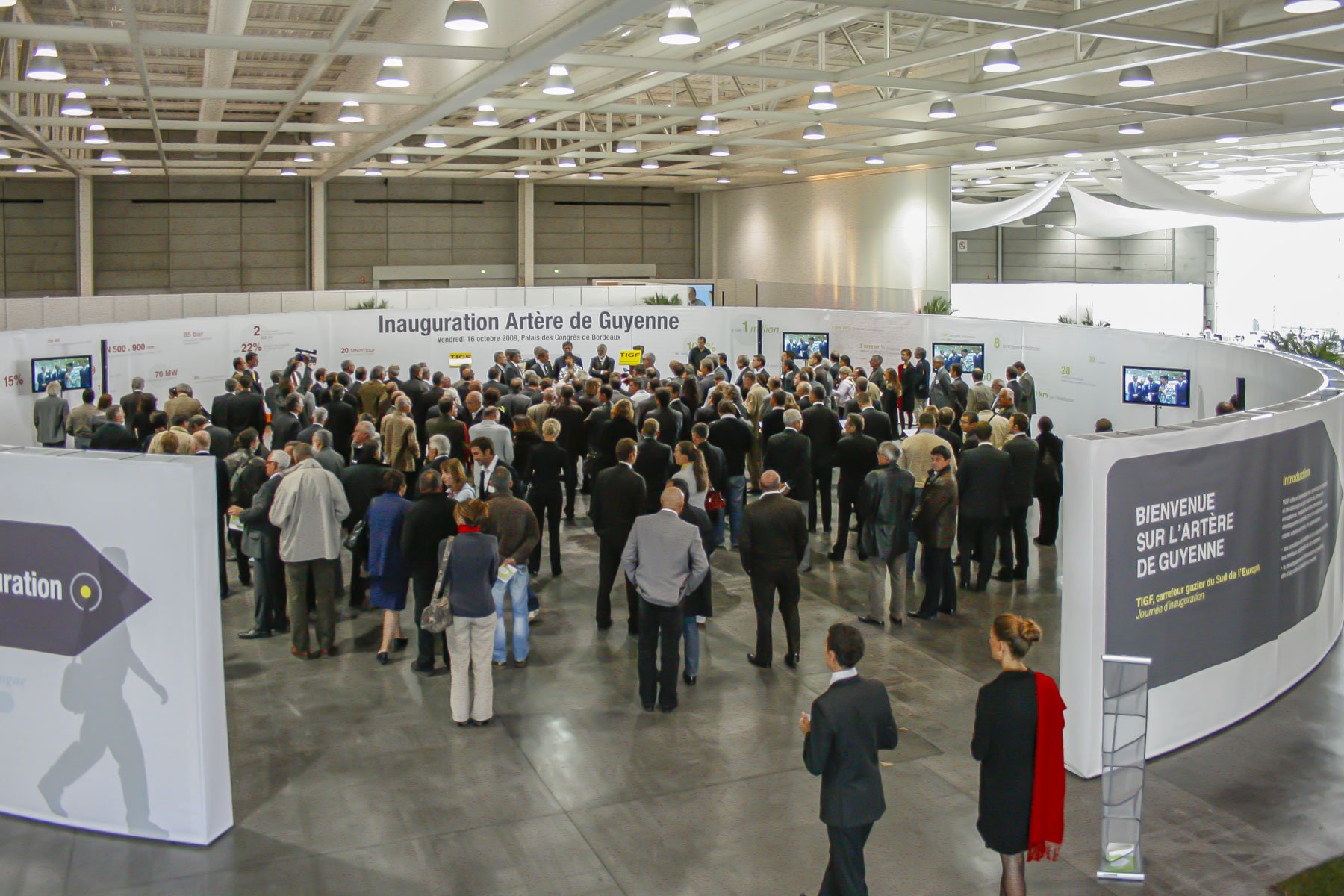 Scénographie d'exposition en intérieur. Vue d'une cérémonie d'inauguration avec de nombreuses personnes se trouvant au centre de l'installation, avec un panneau indiquant « Inauguration Artère de Guyenne ».