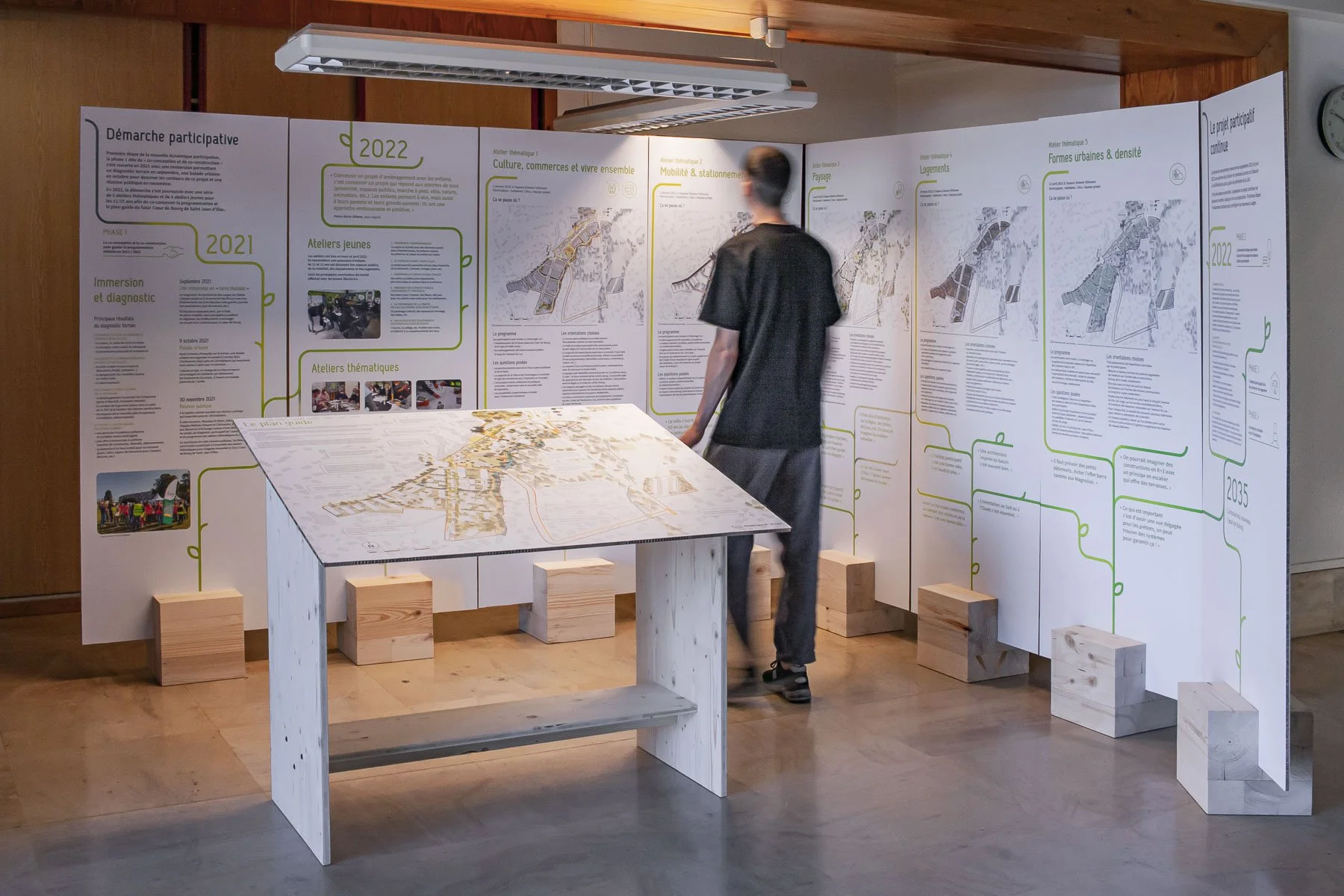 Scénographie d'exposition. Exposition avec de grands panneaux d'information insérés dans des plots piétements en bois clair, un homme regarde les panneaux, une table centrale avec une carte imprimée sur un panneau incliné.