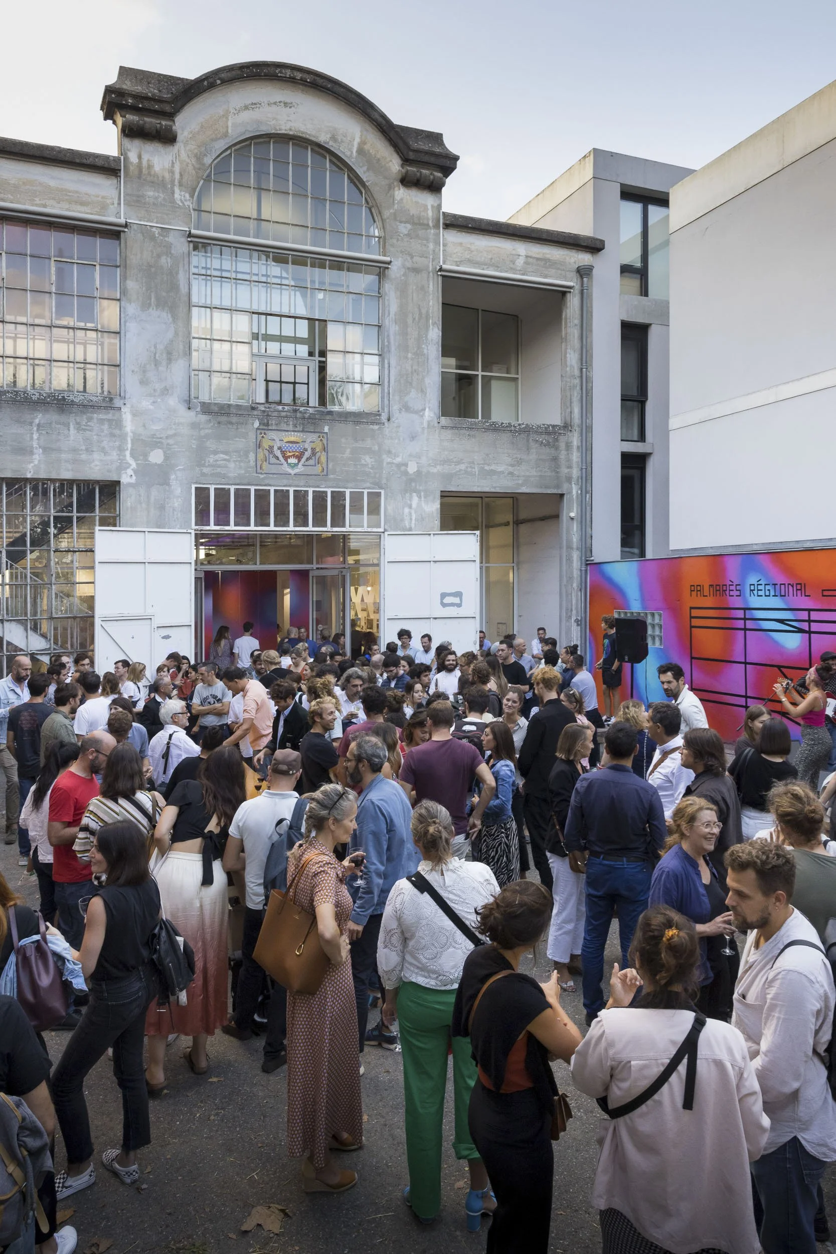 Scénographie d'exposition. Une foule de personnes rassemblée dans un moment festif, en extérieur devant un bâtiment industriel, à l'occasion d'un événement culturel pour le vernissage d'une exposition sur le thème de l'architecture.