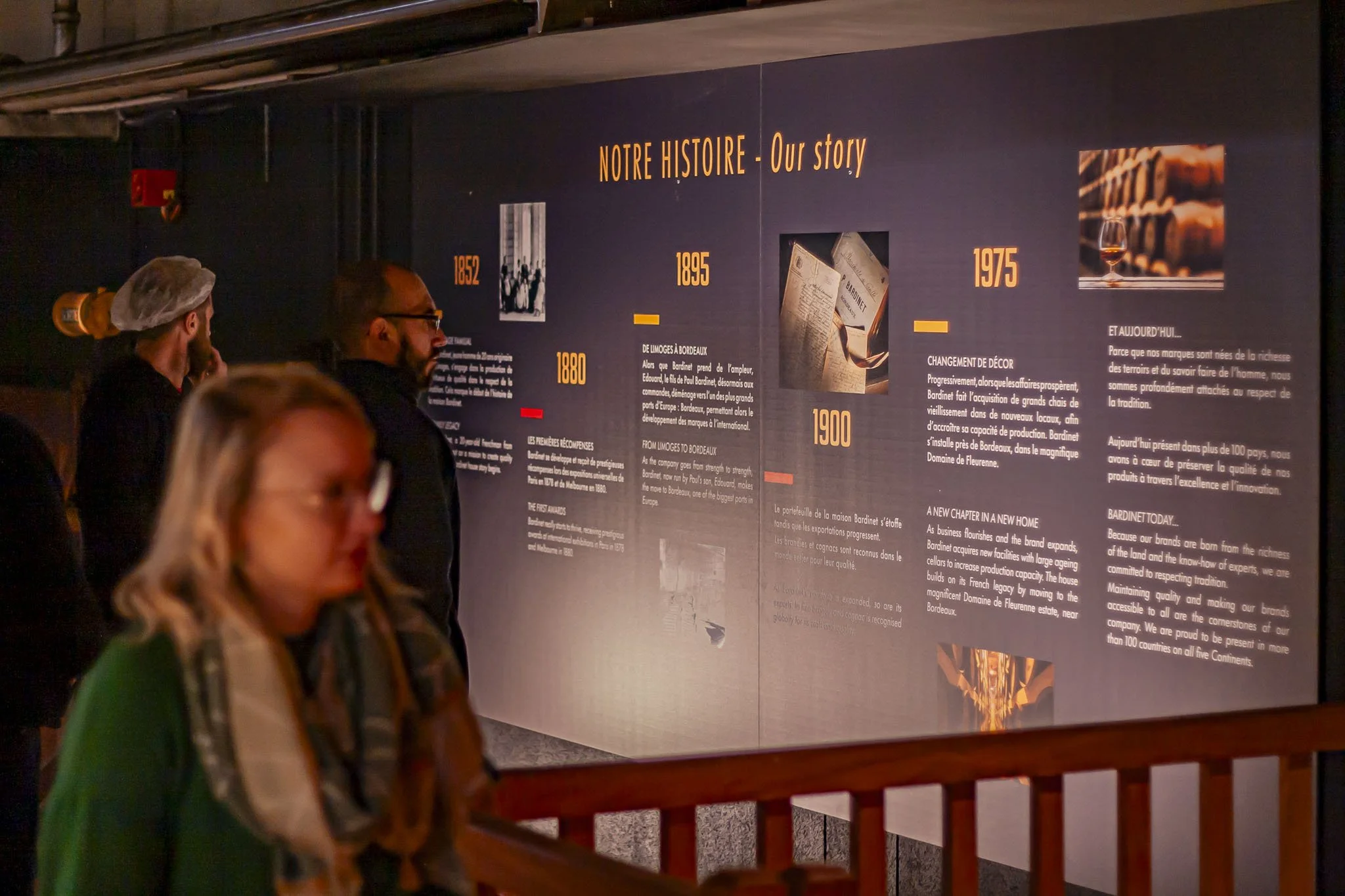 Scénographie d'exposition, valorisation du savoir faire d'entreprise. Intérieur d'un chai à spiritueux. Visiteurs observant une grande frise chronologique sur l'histoire de l'entreprise, depuis sa création à nos jours.