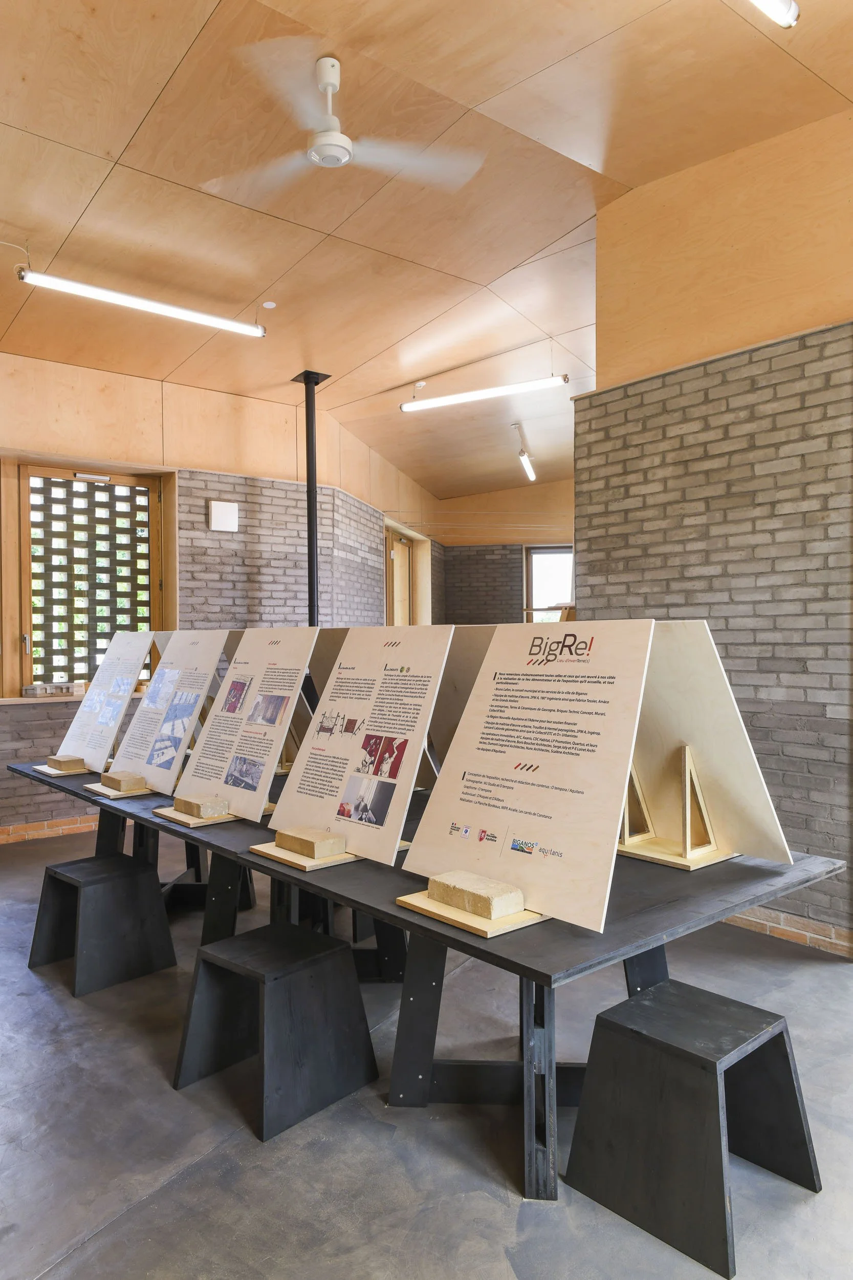 Scénographie d'exposition. Vue intérieure montrant des panneaux bois clair disposés sur des tables en bois huilé teinte ébène. Dans une pièce avec plafond en bois clair, murs en briques grises, et grandes fenêtres.
