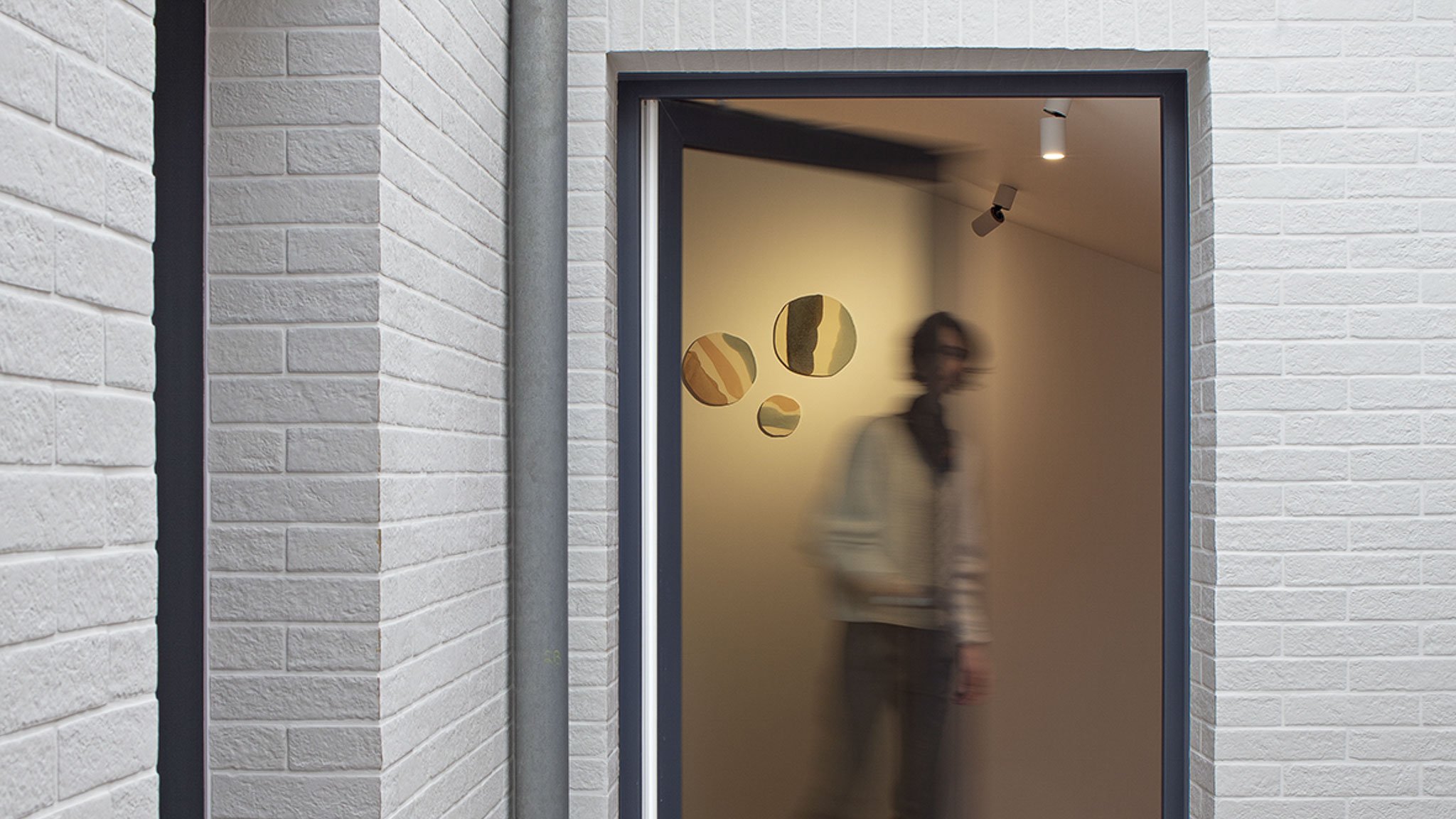 Identité des lieux, transformation d'un internat en hôtel. Détail vue du patio, avec silhouette ouvrant une baie vitrée, et des pièces colorées en céramiques disposées sur le mur derrière elle.