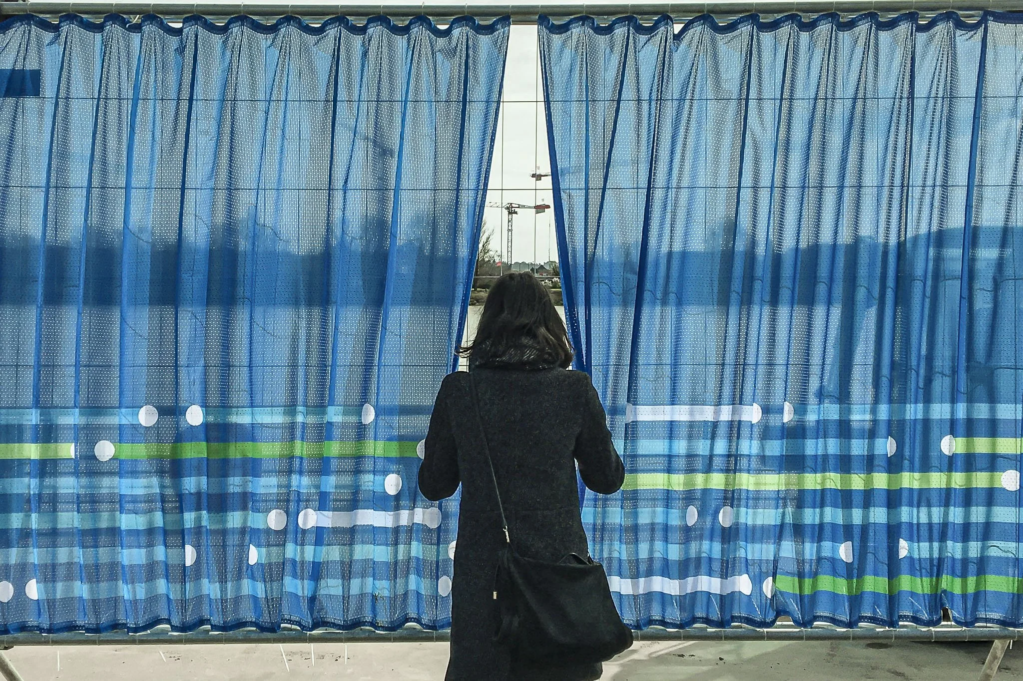 Scénographie en extérieur pour un chantier dans l'espace public. Une femme vue de dos, portant un manteau noir et une sacoche, regarde avec curiosité le chantier à travers une ouverture réalisée dans la bâche de la clôture du chantier.
