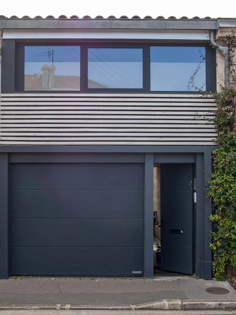 Façade de l'agence avec porte de garage grise et une petite porte de service entrouverte, rangée de fenêtres au premier étage, et une végétation grimpante sur le côté droit.