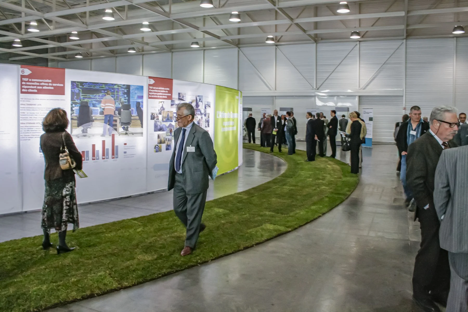 Scénographie d'exposition. Inauguration avec des professionnels, des personnes discutant et observant des affiches d'informations, un parcours de visite végétal avec du vrai gazon est disposé au sol.