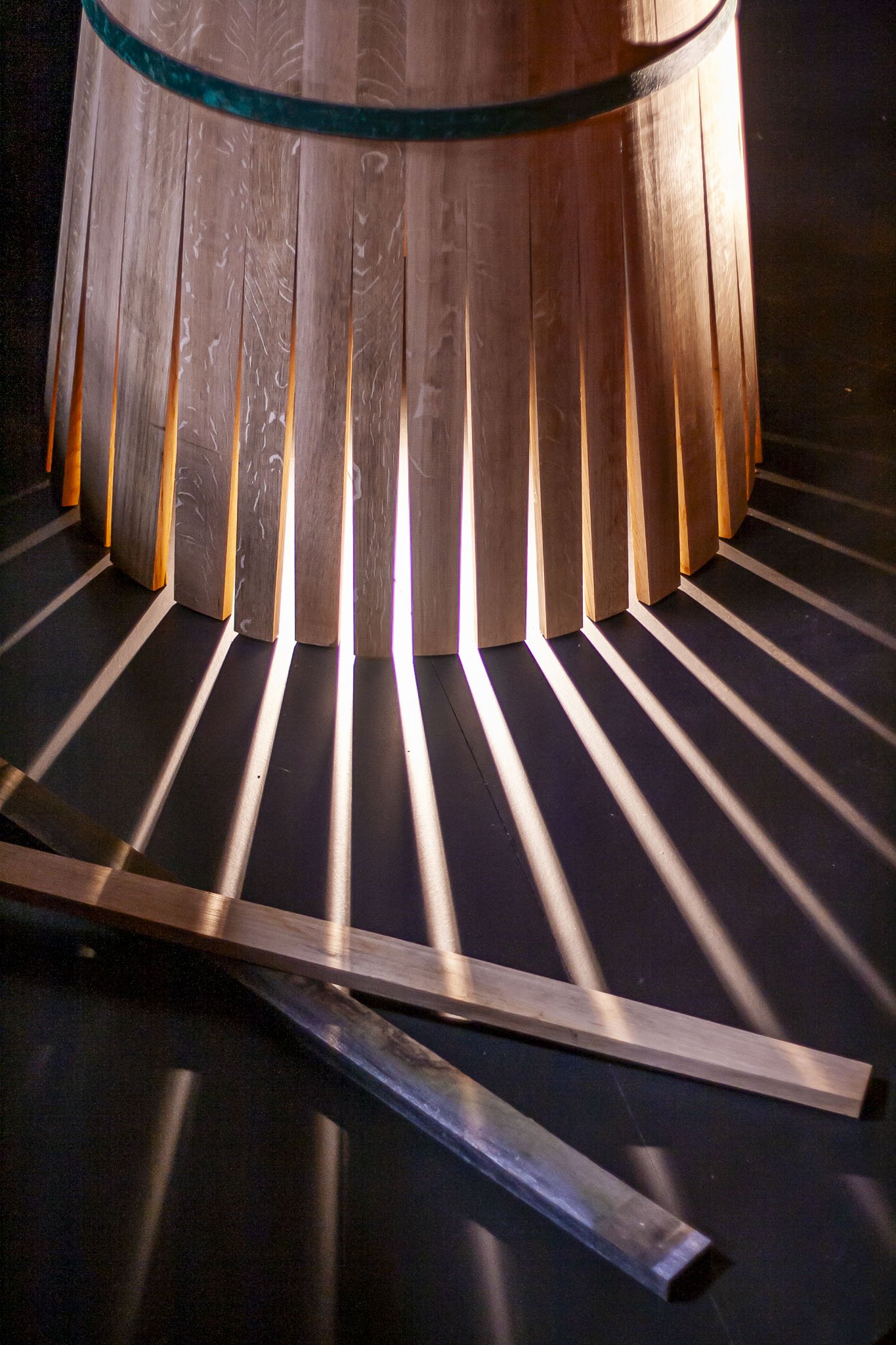 Scénographie d'exposition, valorisation du savoir faire d'entreprise. Intérieur d'un chai à spiritueux. Détail des lames d'une barrique en bois qui laisse passer la lumière à travers des fentes, projetant des rayons lumineux sur un sol sombre.
