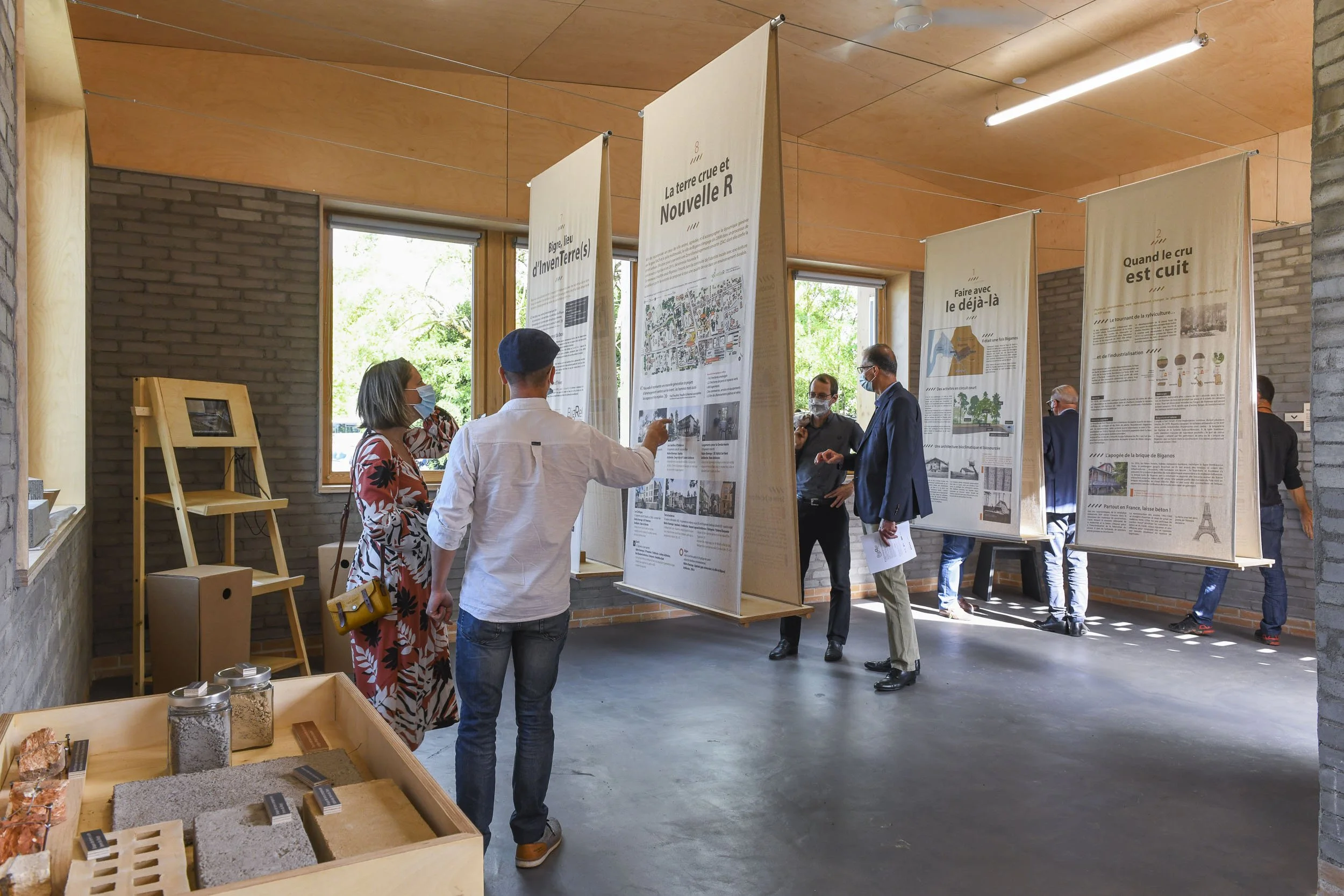 Scénographie d'exposition. Exposition intérieure avec plusieurs panneaux d'information suspendus, quelques visiteurs discutant, un présentoir avec des échantillons en avant-plan, murs en pierre et fenêtres laissant entrer la lumière naturelle.