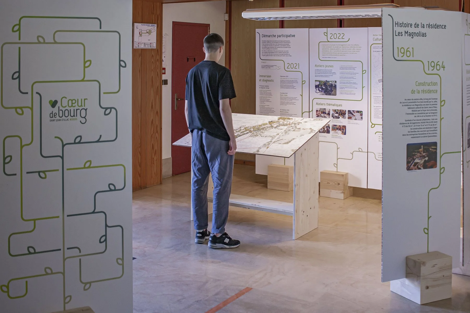 Scénographie d'exposition. Un jeune homme regarde un panneau d'exposition avec des textes et des images. La scène se déroule dans une salle d'exposition avec un sol en bois clair, des panneaux d'affichage blancs et un mur en bois en arrière-plan.