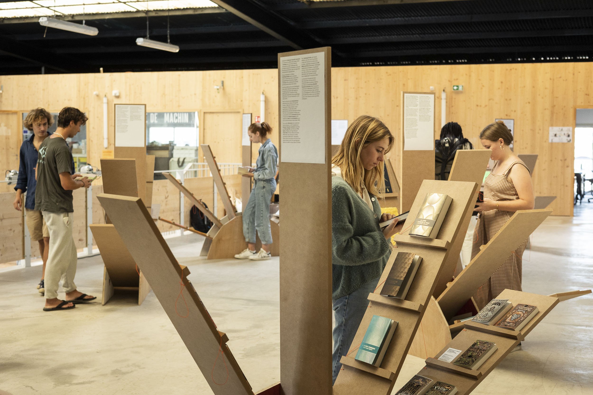 Scénographie d'exposition en intérieur, présentant des livres graphiques. Plusieurs personnes lisant et regardant des livres à côté des mobiliers scéniques d'exposition, des grands modules en bois en forme d'éventail.