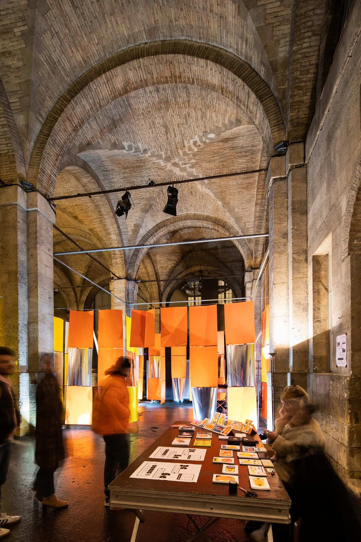 Scénographie d'exposition en intérieur, présentant des projets graphiques, un espace avec voûtes en pierre, comprenant une grande installation de panneaux oranges suspendus et un bureau avec des brochures et dépliants.