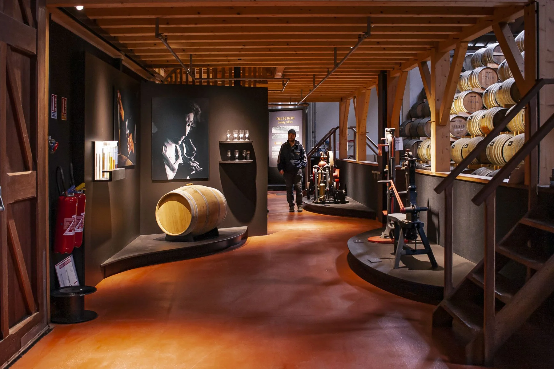 Scénographie d'exposition. Intérieur d'un chai à spiritueux, avec des tonneaux en chêne, du contenu d'exposition, des grandes photos, des vieux outils, des plateformes en bois peintes en noir avec des courbes, au sol, et une personne se promenant.