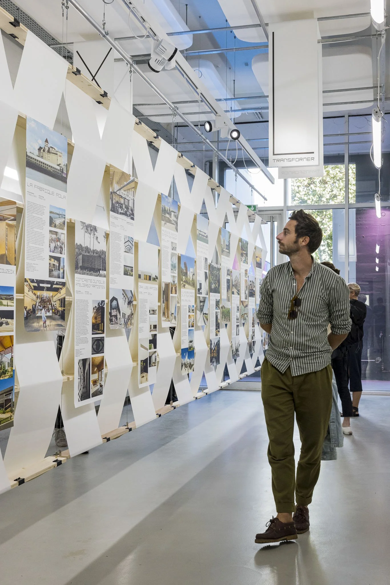 Scénographie d'exposition. Un homme regarde une exposition d'images et de textes dans une galerie moderne, avec une structure en bois et des affiches suspendues, en intérieur lumineux avec de larges fenêtres.
