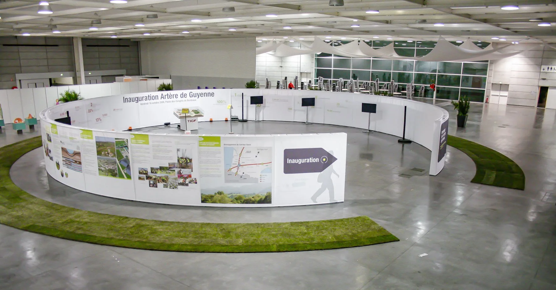 Scénographie d'exposition en intérieur. Aperçu du dispositif d'exposition avec une longue paroi verticale disposée au sol en ellipse. Un sol en béton, quelques plantes en pot, un parcours de visite en gazon, et des chaises empilées dans le fond.