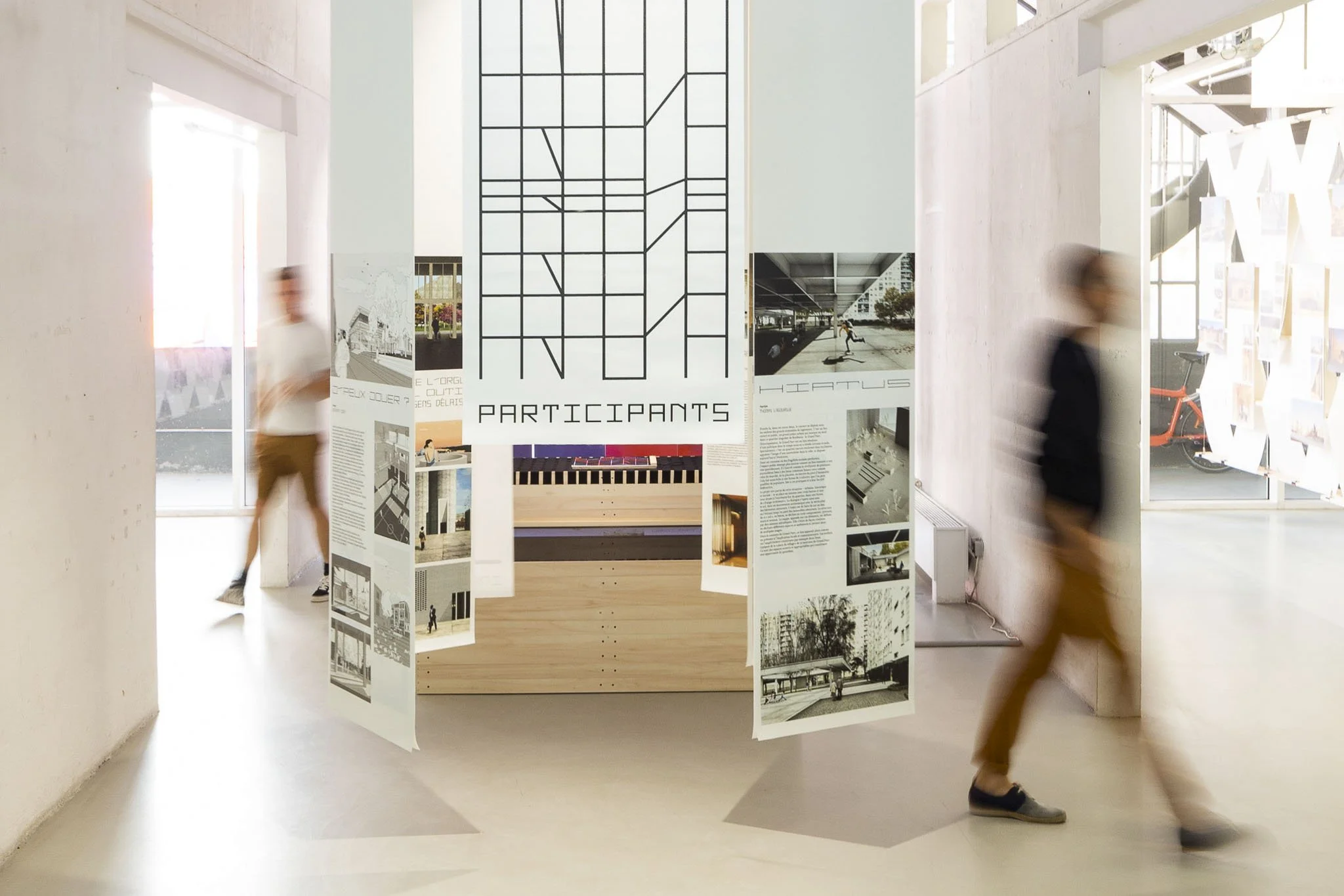 Scénographie d'exposition. Exposition intérieure avec panneaux d'affichage, personnes floues en mouvement, grande œuvre graphique avec le mot "PARTICIPANTS" suspendue au centre, et espace moderne lumineux avec grandes fenêtres.