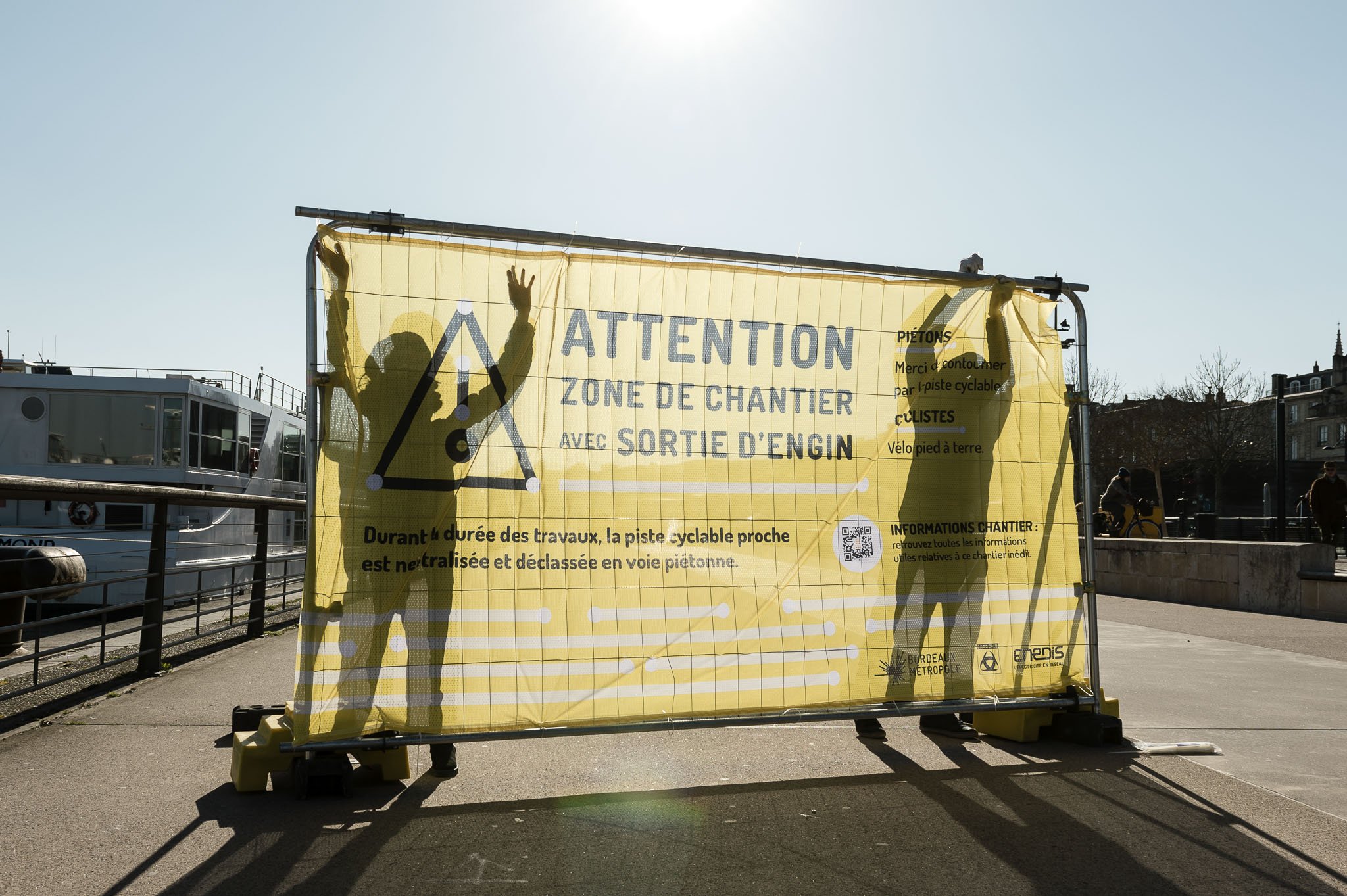 Scénographie en extérieur pour un chantier dans l'espace public. Grande bâche de chantier jaune avec silhouettes de deux personnes et texte d'avertissement en français, indiquant une zone de chantier avec sortie d'engin.
