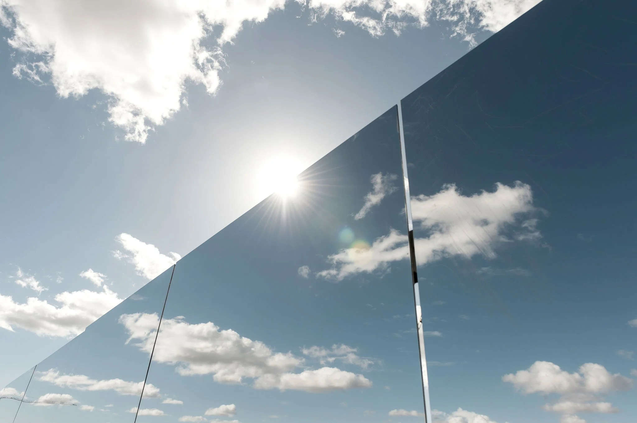 Scénographie en extérieur pour un chantier dans l'espace public. Des panneaux miroirs réfléchissant le ciel et les nuages avec le soleil brillant derrière, créant un effet de reflet