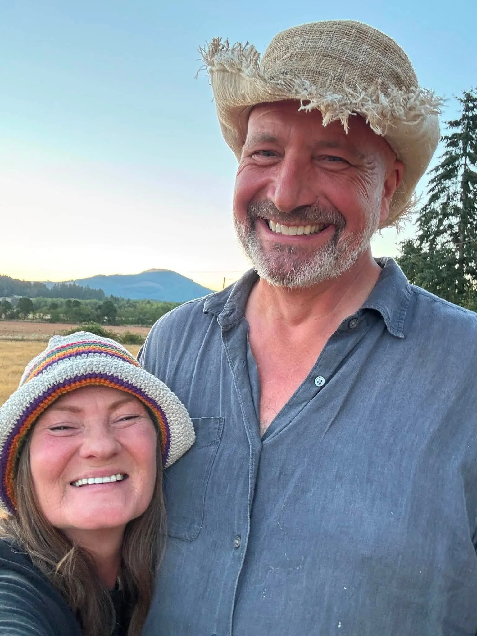 A man with a partly gray beard wearing a light-colored woven cowboy hat and a blue button-up shirt, smiling outdoors with a landscape of trees, hills, and a blue sky in the background.