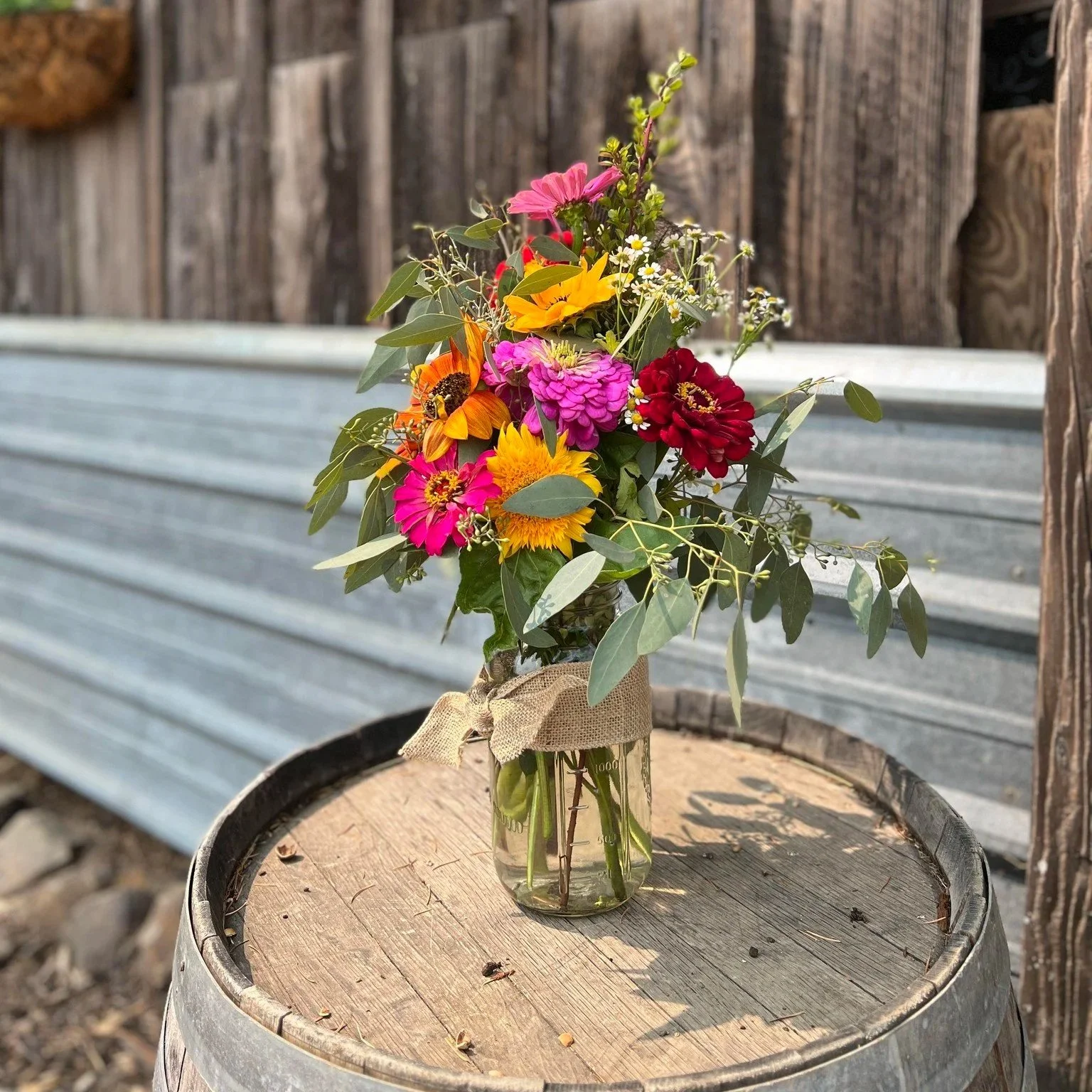 A bouquet of colorful flowers in a glass jar, tied with a burlap ribbon, placed on a wooden barrel outdoors.