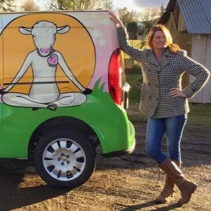 A woman leaning against a colorful vehicle featuring a cartoon cow in a meditation pose, outdoors on a sunny day.