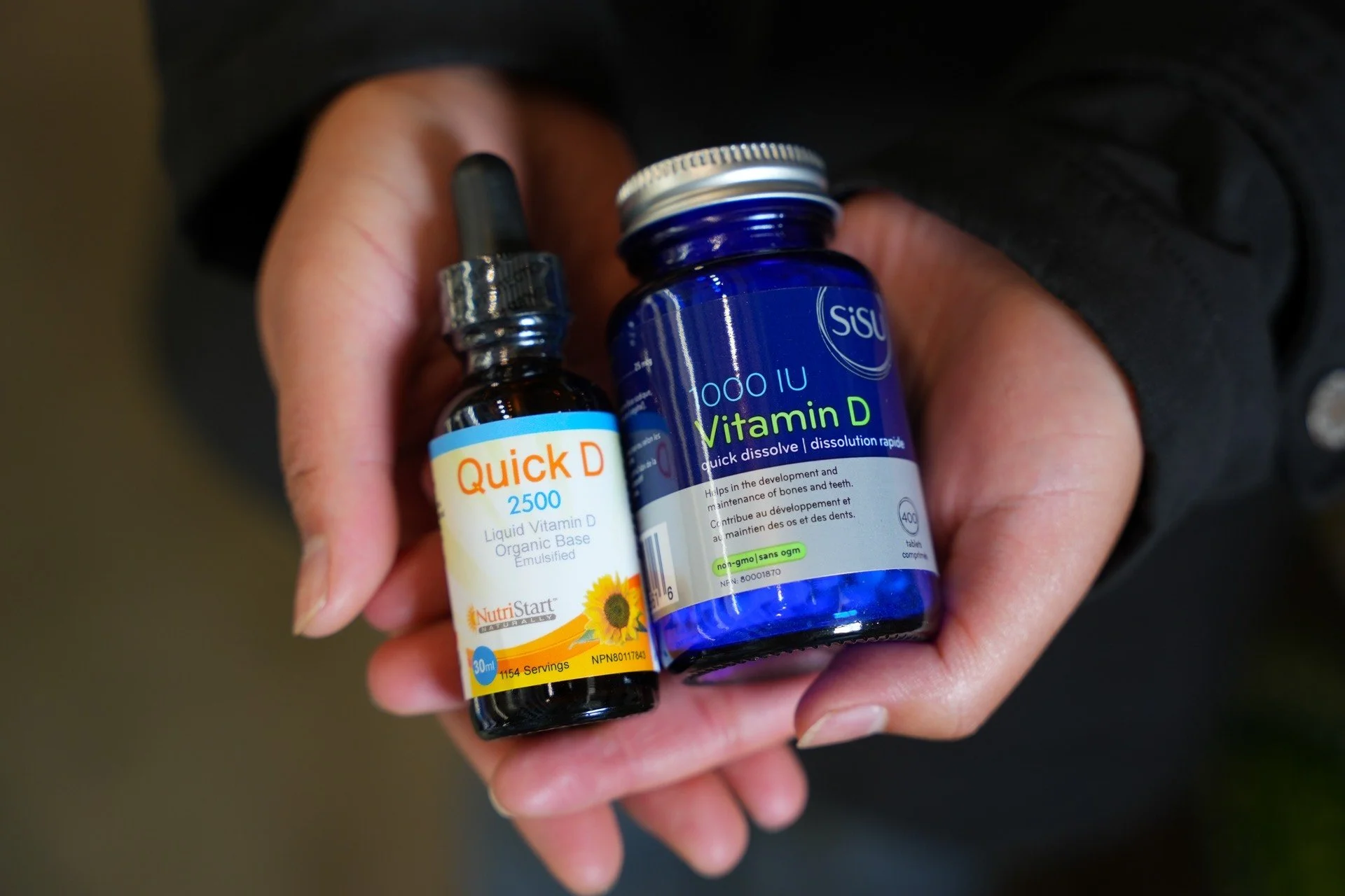 A person holding two bottles of dietary supplements, one labeled 'Quick D 2500' and the other 'Vitamin D 10000 IU', both in their palm with the person's fingers visible on the sides.