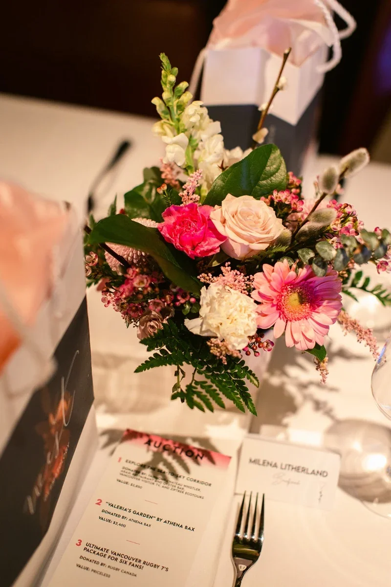 A floral centerpiece with pink, white, and peach flowers on a table at an event, with auction and place cards nearby.