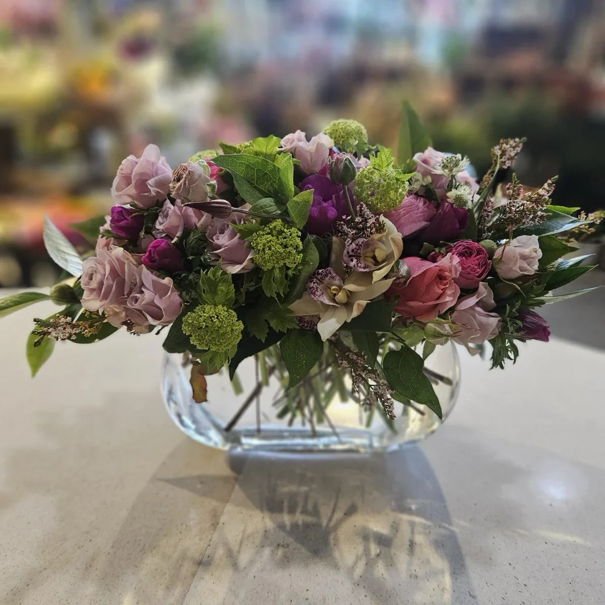 A colorful bouquet of various flowers including pink roses, white orchids, purple ranunculus, and green foliage in a clear glass vase on a white surface.