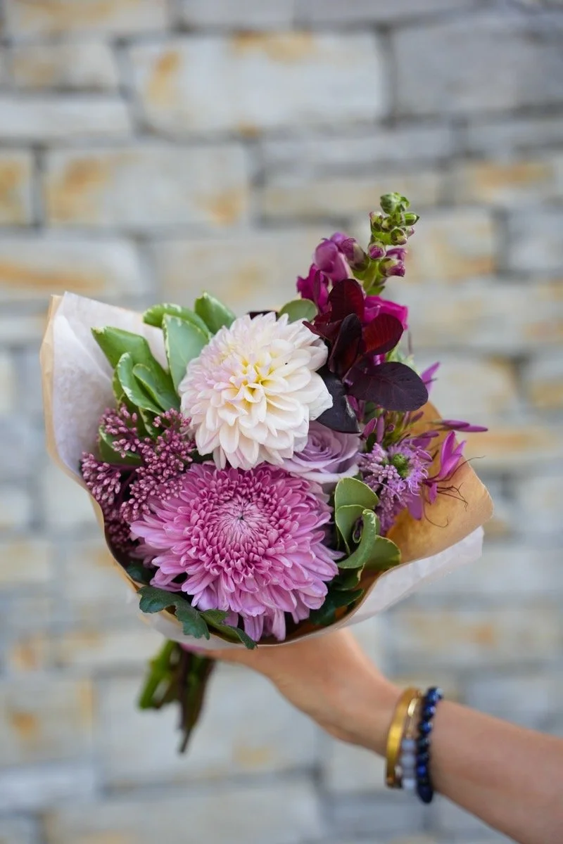 A bouquet of pink and white flowers, held by a hand with bracelets, against a brick wall background.