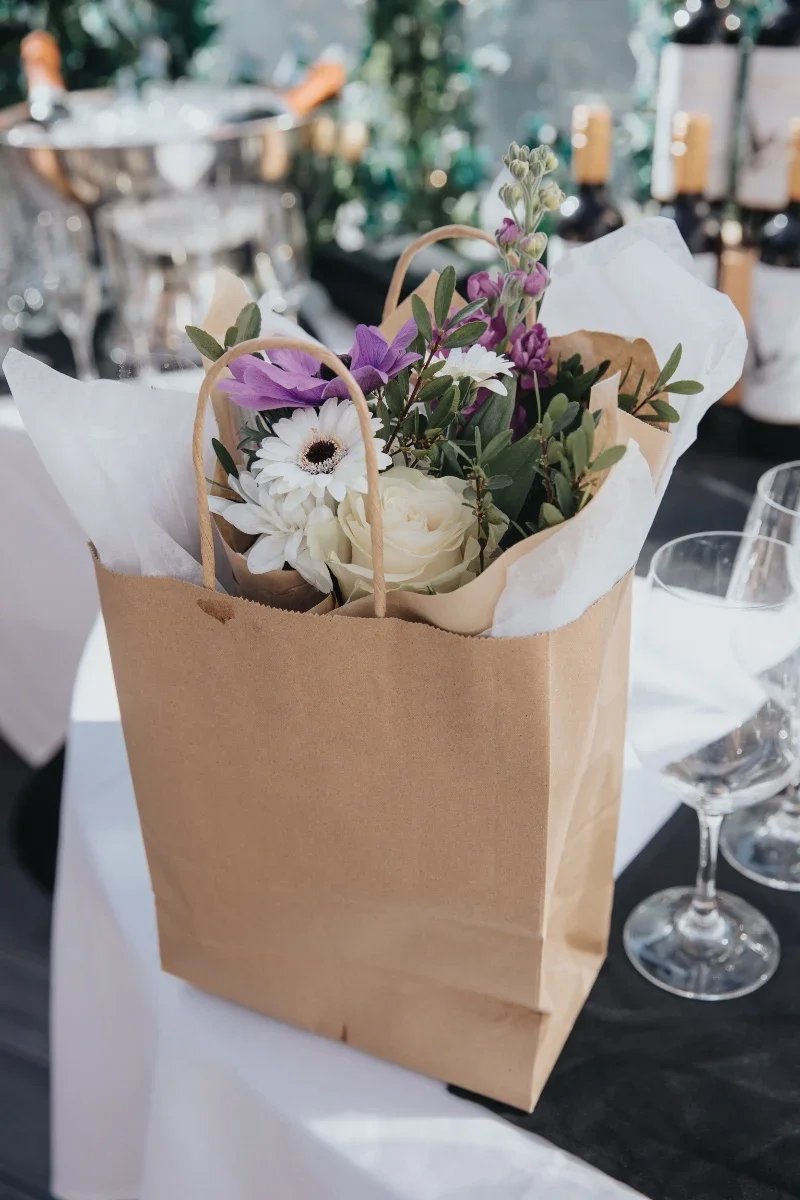 A brown paper gift bag filled with flowers, including white roses, purple bellflowers, and white daisies, placed on a table with glassware around in a setting that appears to be a restaurant or event.