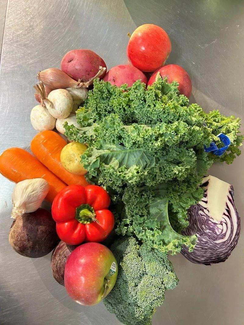 A variety of fresh vegetables and fruits including red apples, radishes, garlic, carrots, a red bell pepper, broccoli, purple cabbage, beets, and kale on a gray surface.
