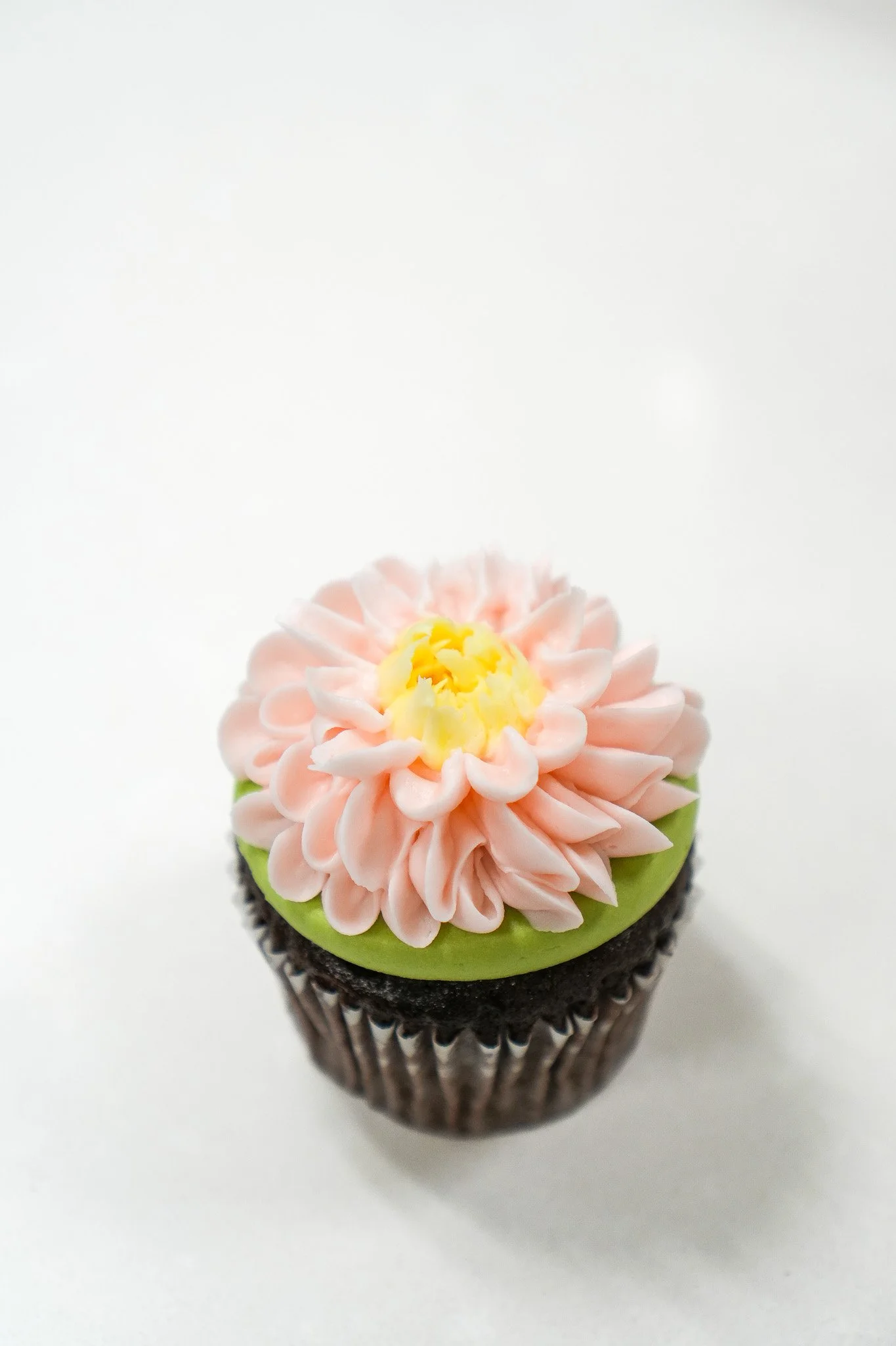 Decorated cupcake with pink flower frosting and green fondant on top, in a paper cupcake liner, on a white background.