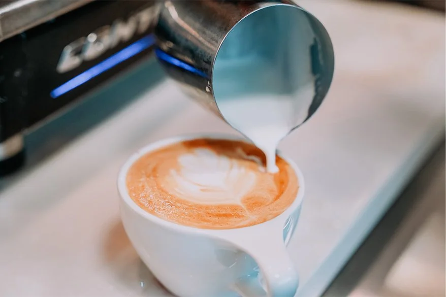 A barista pours steamed milk into a cup of espresso, creating latte art on top.