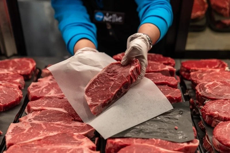 Person wearing gloves placing a raw steak on a tray with more steaks around.