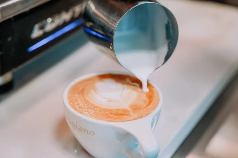 A stainless steel milk pitcher pouring steamed milk into a cup of espresso, creating latte art on top of a latte.