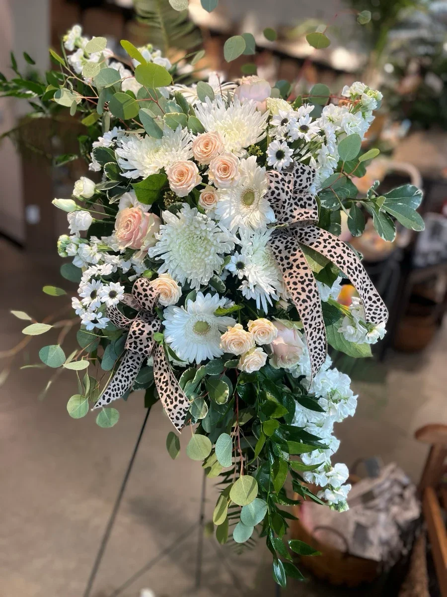 A floral arrangement with white and pale pink roses, white daisies, white chrysanthemums, and greenery, including eucalyptus leaves, decorated with leopard print ribbons.
